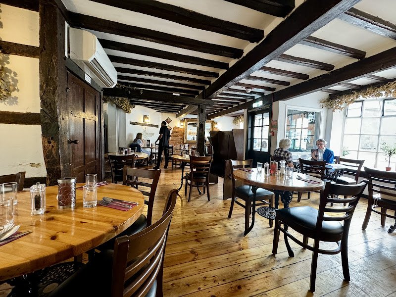 Interior of a cozy restaurant with wooden floors and beams, wooden and upholstered chairs, tables set with glasses and cutlery, and large windows letting in natural light, with a few patrons dining.