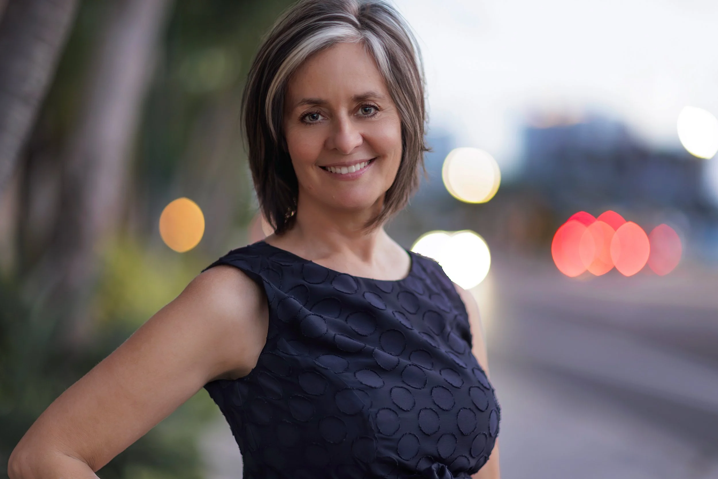 A woman with short, brown hair and blonde highlights, smiling outdoors in the evening, wearing a sleeveless navy blue dress with a textured pattern, standing in front of a blurred background of trees and city lights.