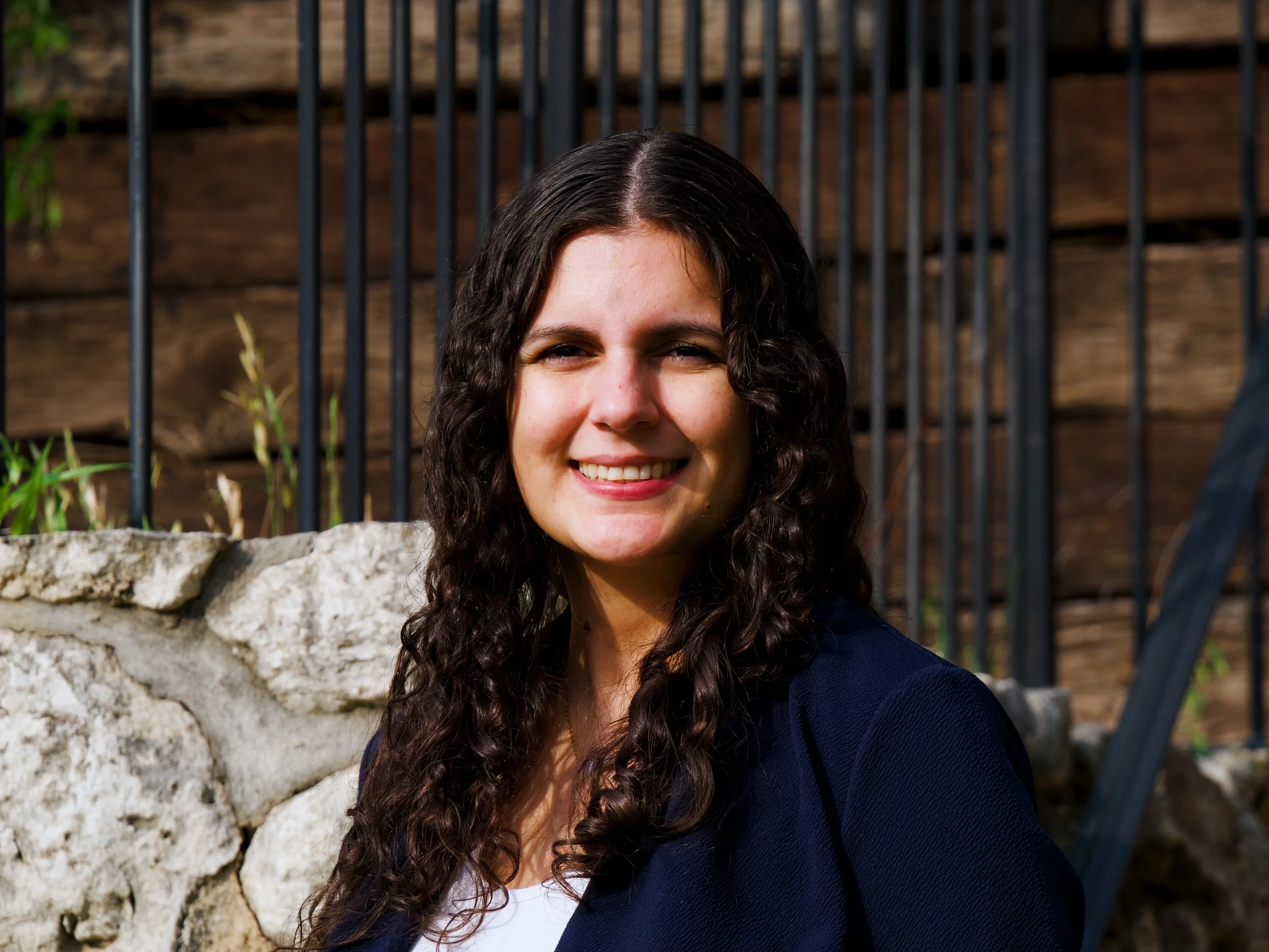 'A woman with long dark curly hair, smiling, wearing a navy blazer and white top, standing outdoors near a stone wall and black metal fence, with a wooden fence in the background.
