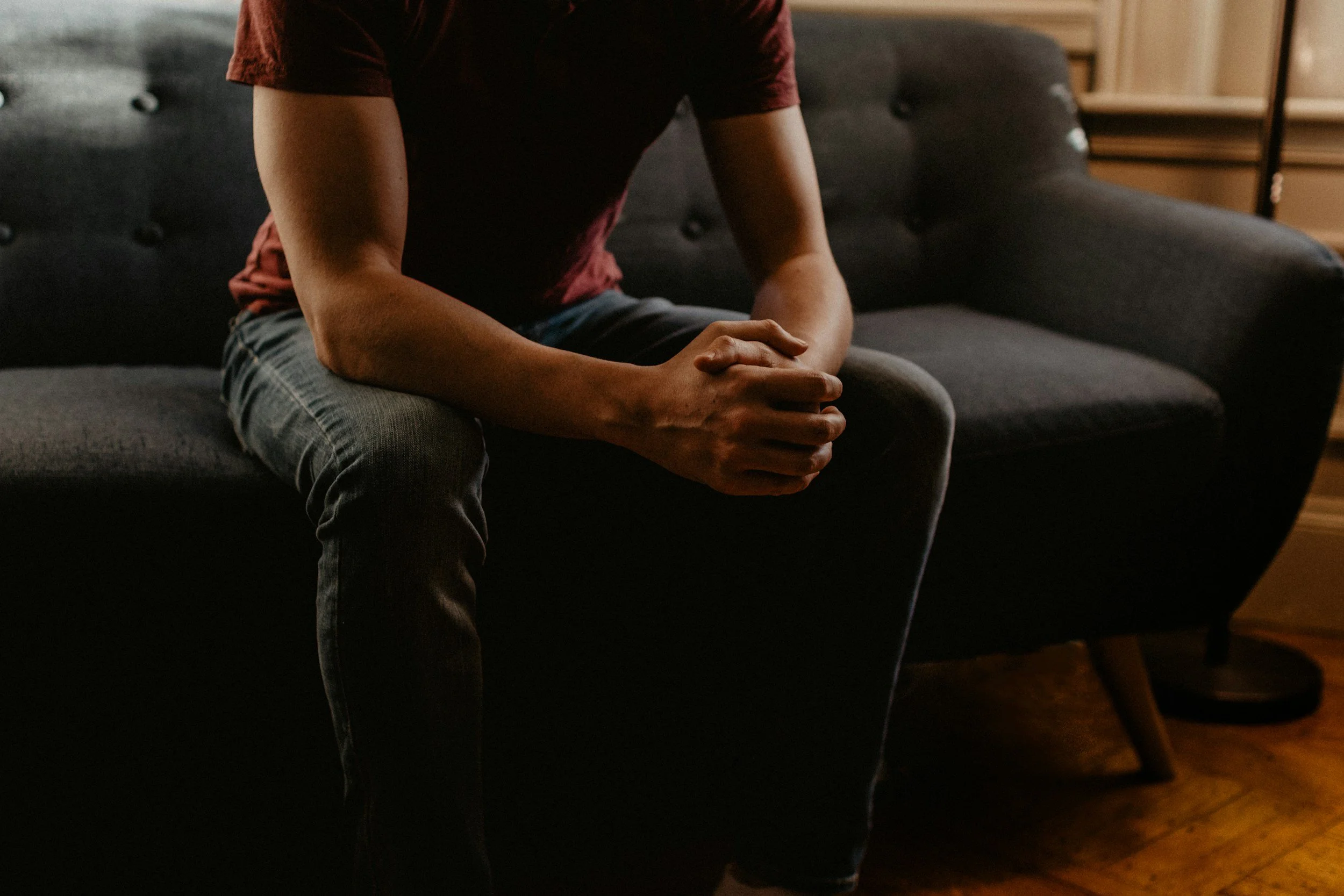 Person sitting on a dark gray sofa with hands clasped and head bowed in a dimly lit room.