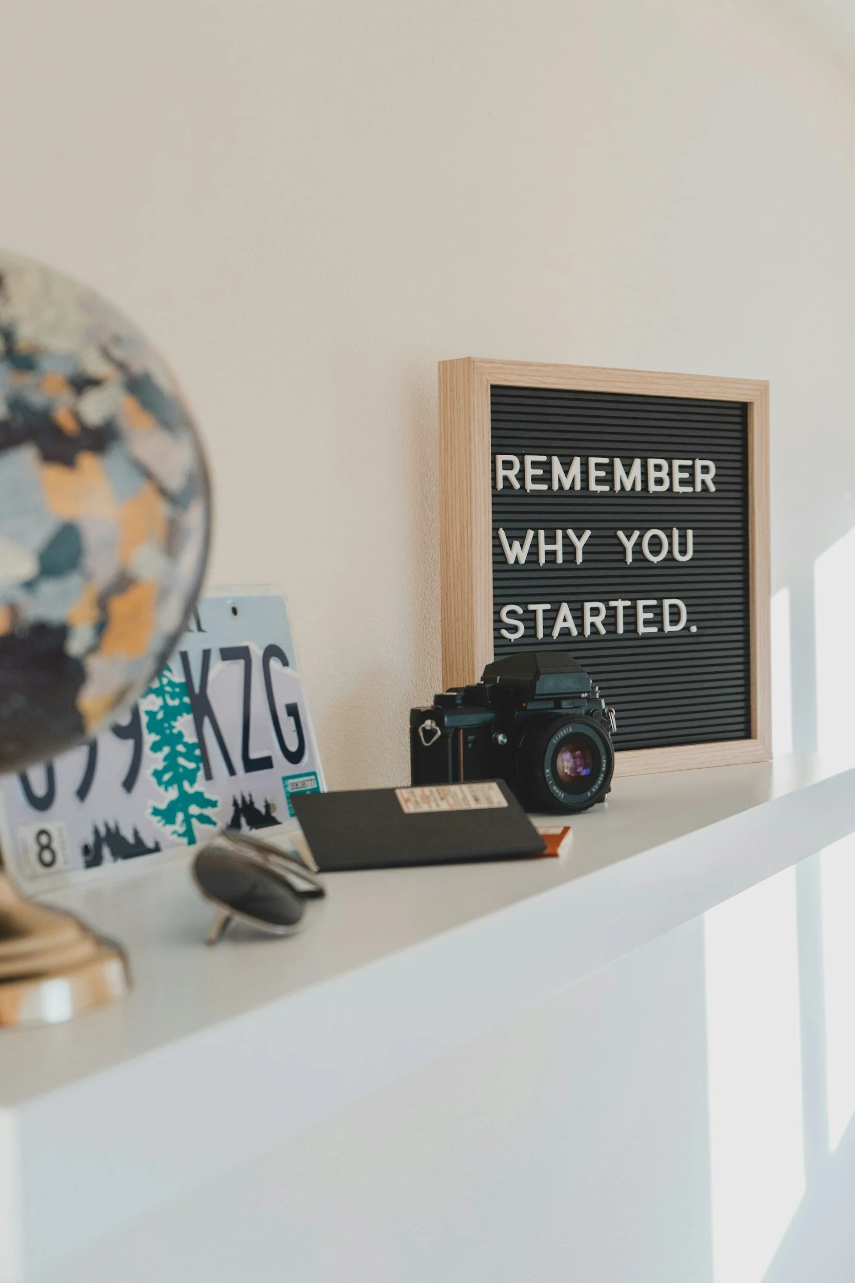 Shelf with globe, car license plate, camera, notebook, glasses, and a motivational quote sign that says, "Remember why you started."