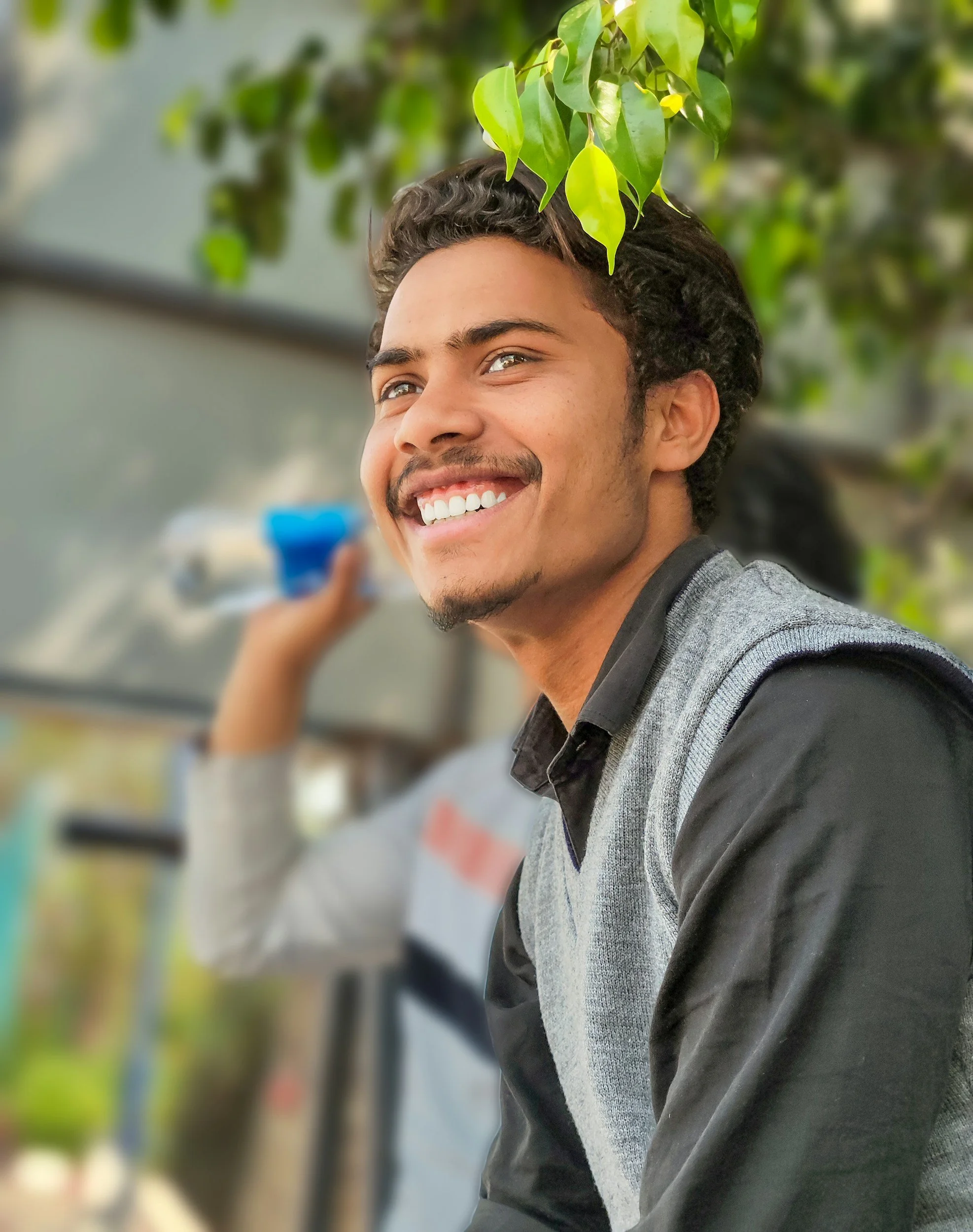 Young man with curly hair and a goatee smiling, sitting outdoors next to a tree, with blurred background of another person and greenery.