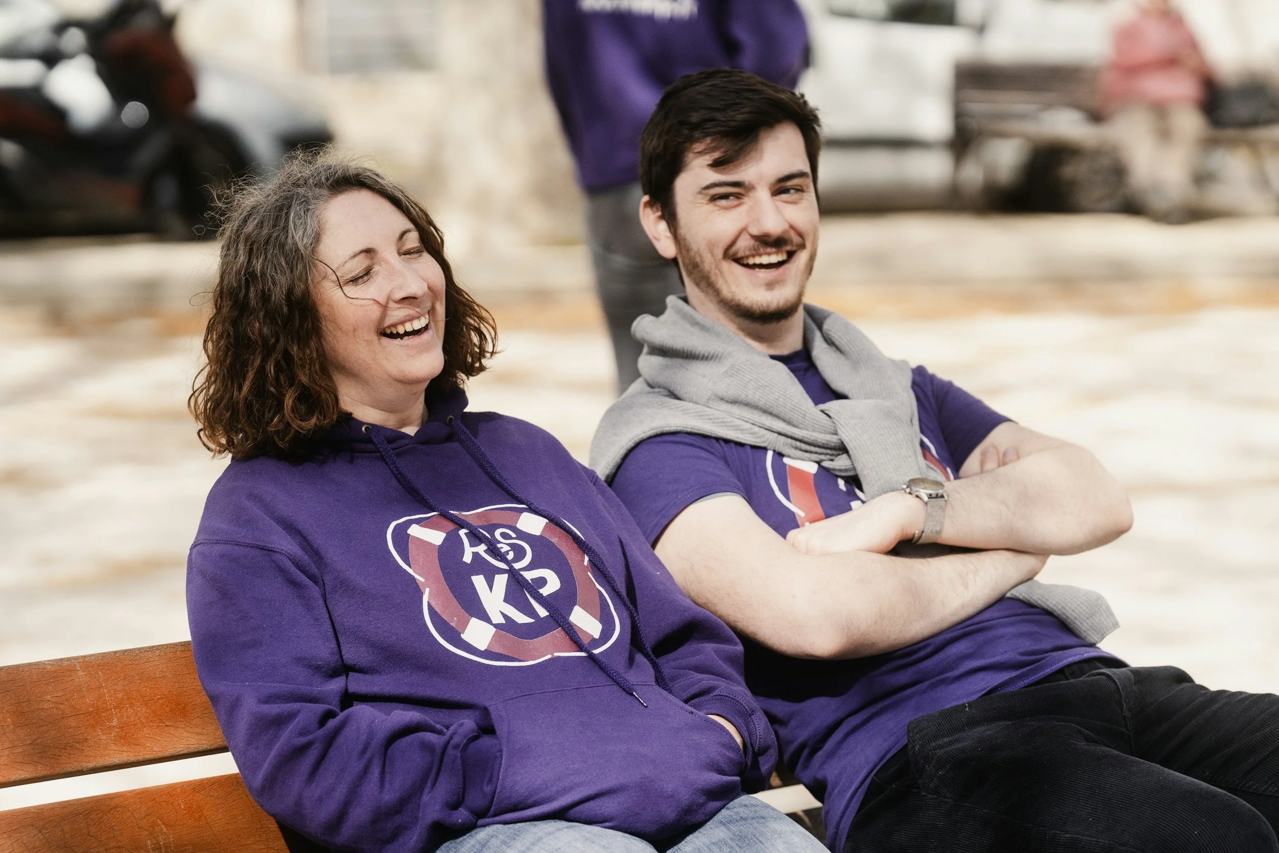 Two people sitting on a park bench, smiling and laughing. They are wearing purple shirts with a logo and have casual attire. The background shows a park setting with some blurred figures and cars.