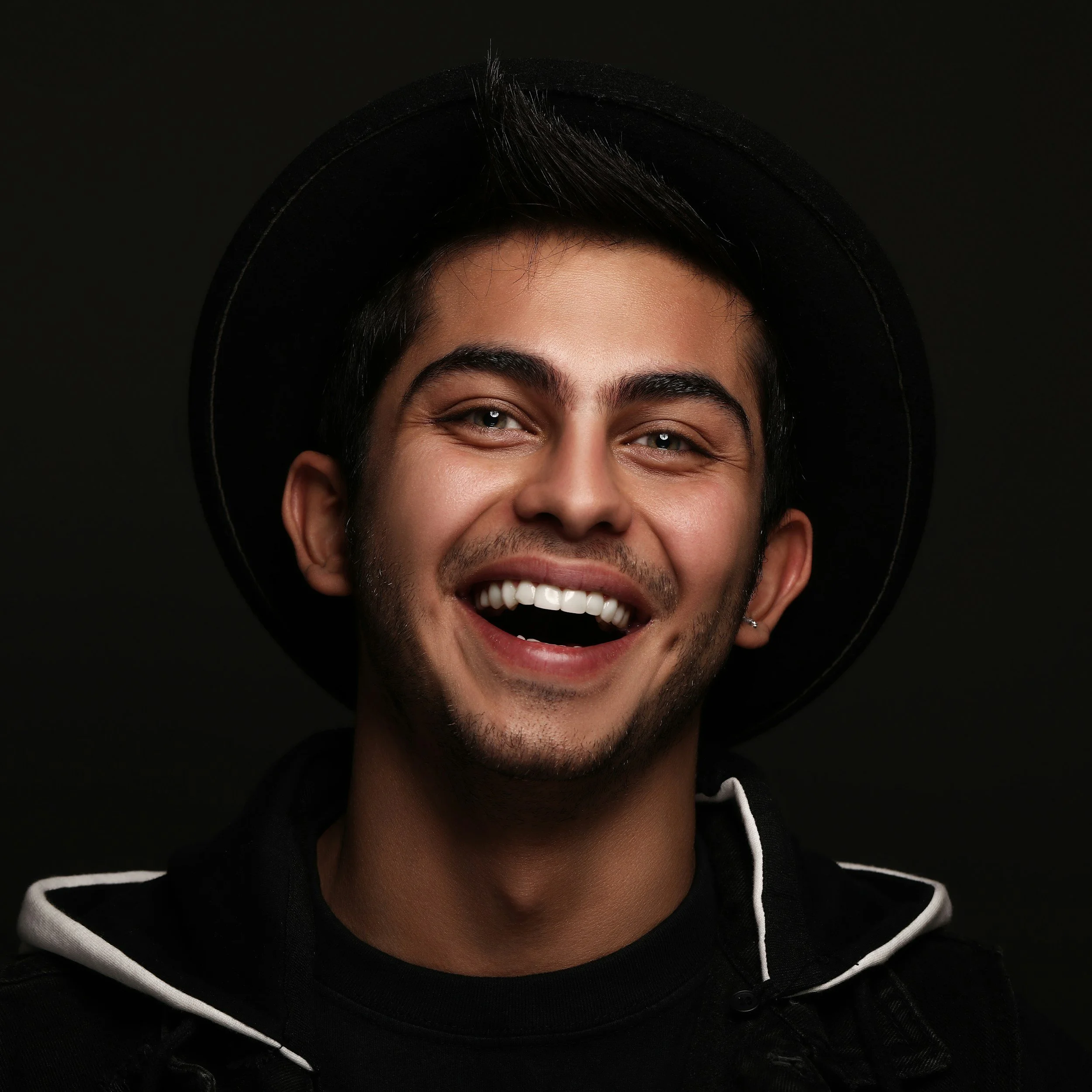 A young man with dark hair, wearing a black fedora hat and a black hoodie, smiling with teeth showing against a dark background.