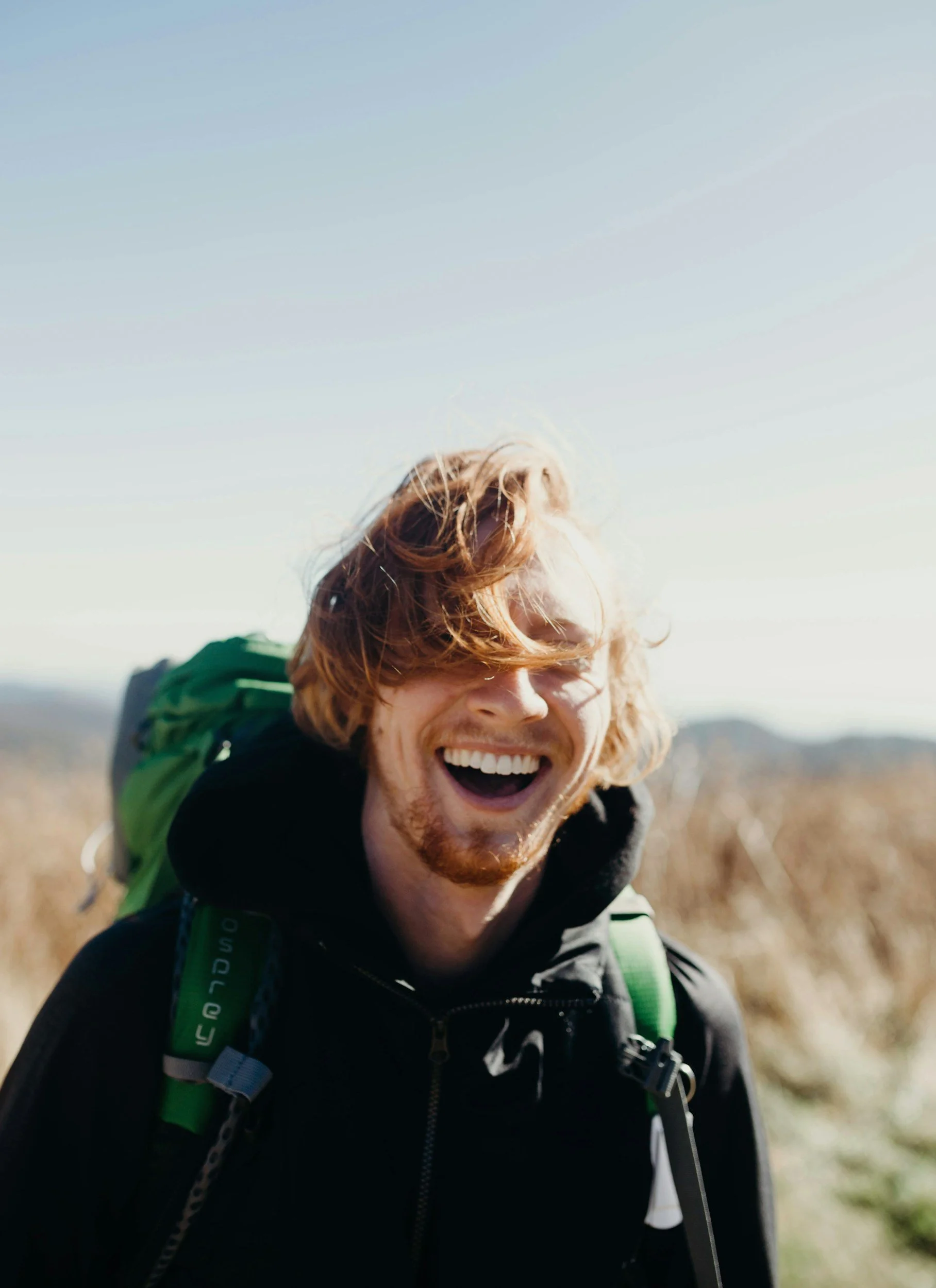 A young man with red hair and a beard smiling outdoors, wearing a black jacket and a green backpack, in a natural setting with dry grass and a clear blue sky.