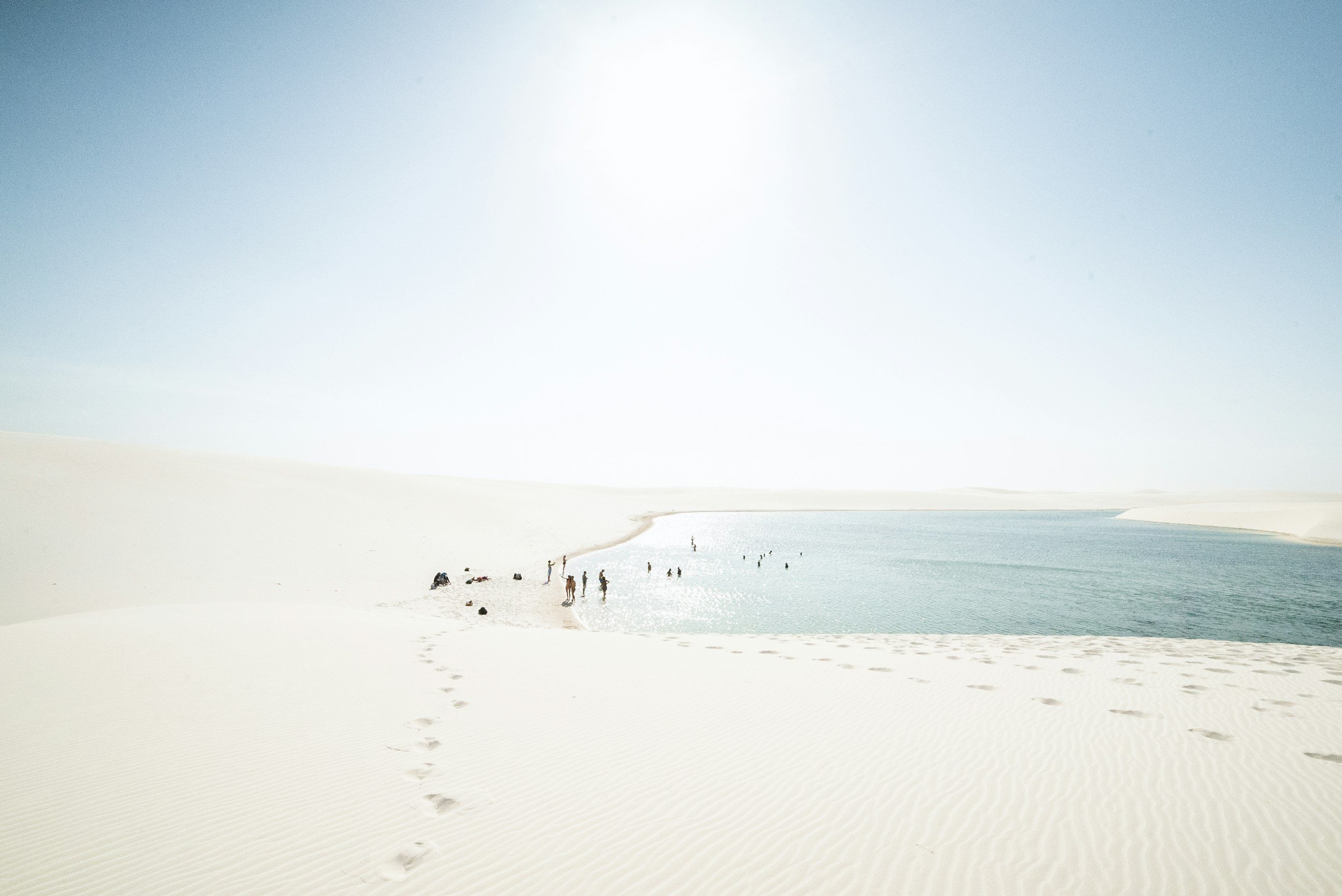 White sand dunes surrounding a blue water lagoon with people swimming and walking along the shore, under a bright sun in a desert landscape.