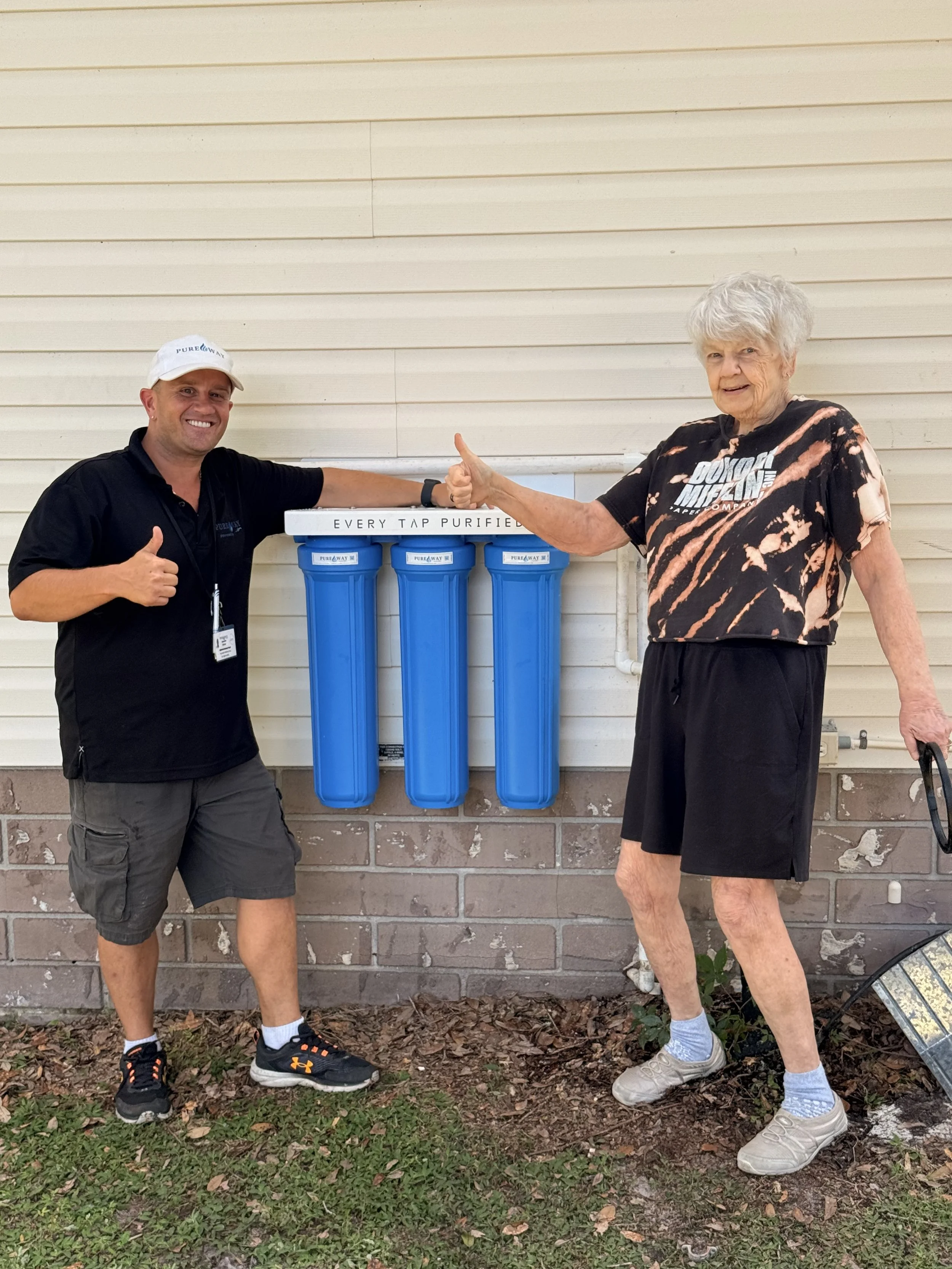 a man and a woman standing outside giving a thumbs up while standing next to a whole house water filter attached to her home