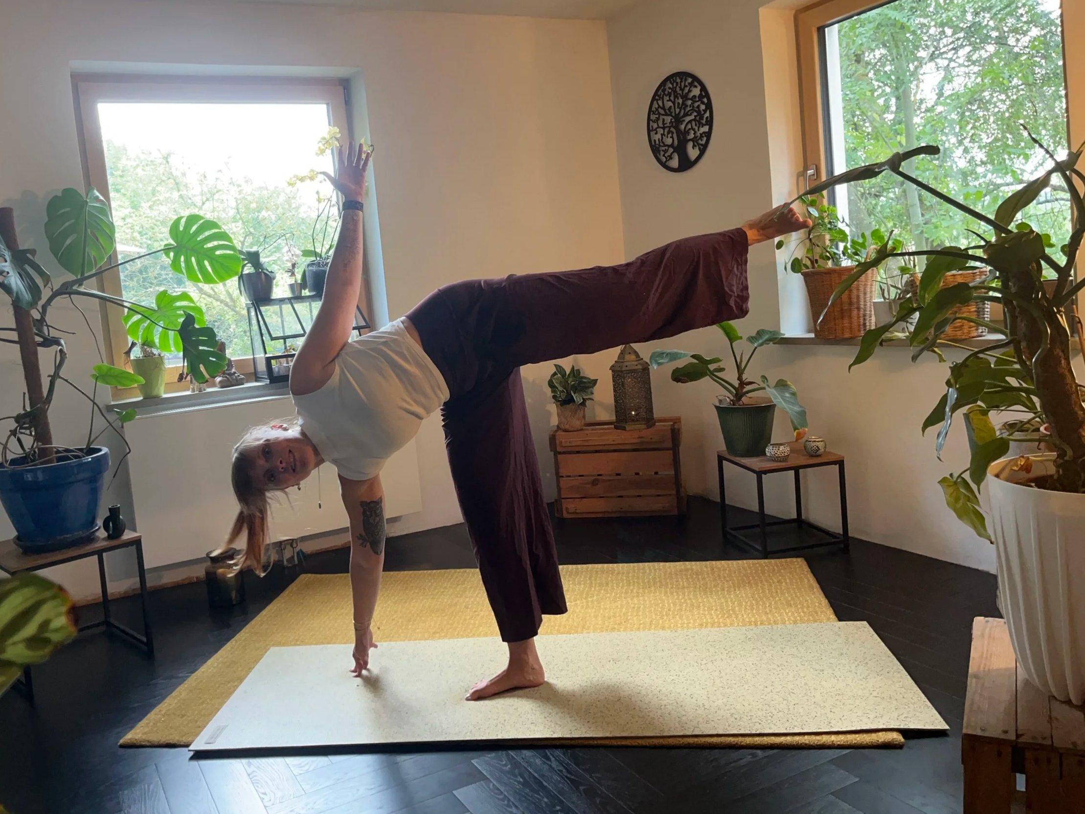 Frau macht Yoga in einem hellen Raum mit großen Fenstern, umgeben von Pflanzen.