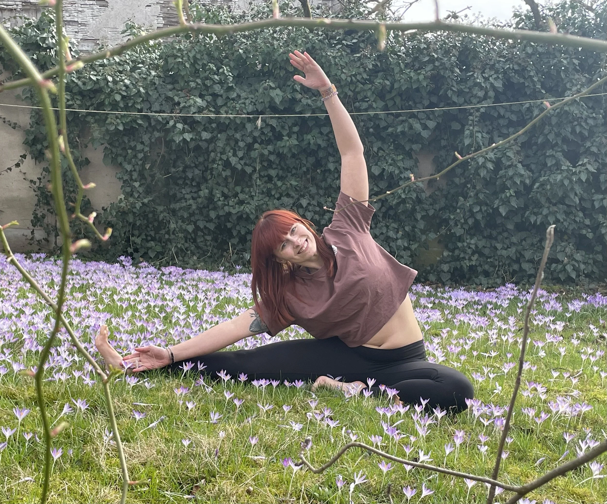 Eine Frau mit roten Haaren macht eine Yogapose auf einer Wiese voller lila Krokusse, umgeben von Hecken und Zweigen.