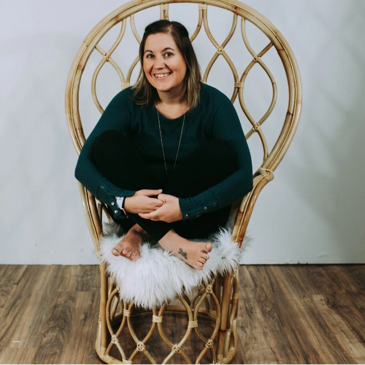 A woman with shoulder-length brown hair smiling, sitting barefoot with her knees drawn up, on a wicker peacock chair with a white furry cushion, against a plain white background.