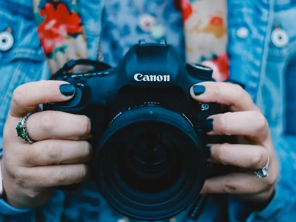 Close-up of a person holding a Canon DSLR camera, wearing rings and painted nails.