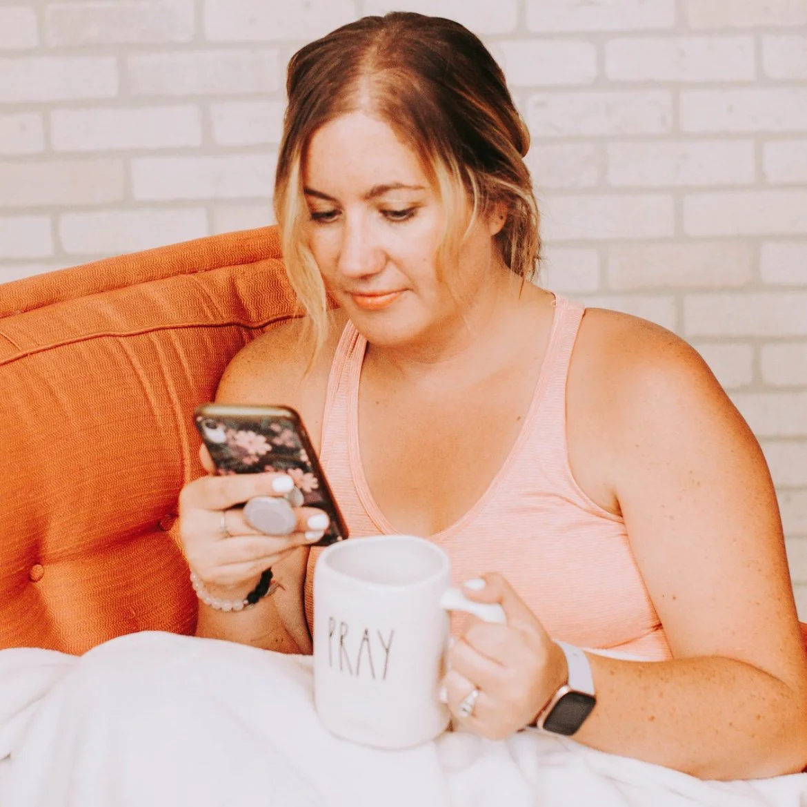 A woman with light skin and shoulder-length hair, wearing a pink tank top, is sitting in bed, looking at her phone while holding a white mug with 'PRAY' written on it.