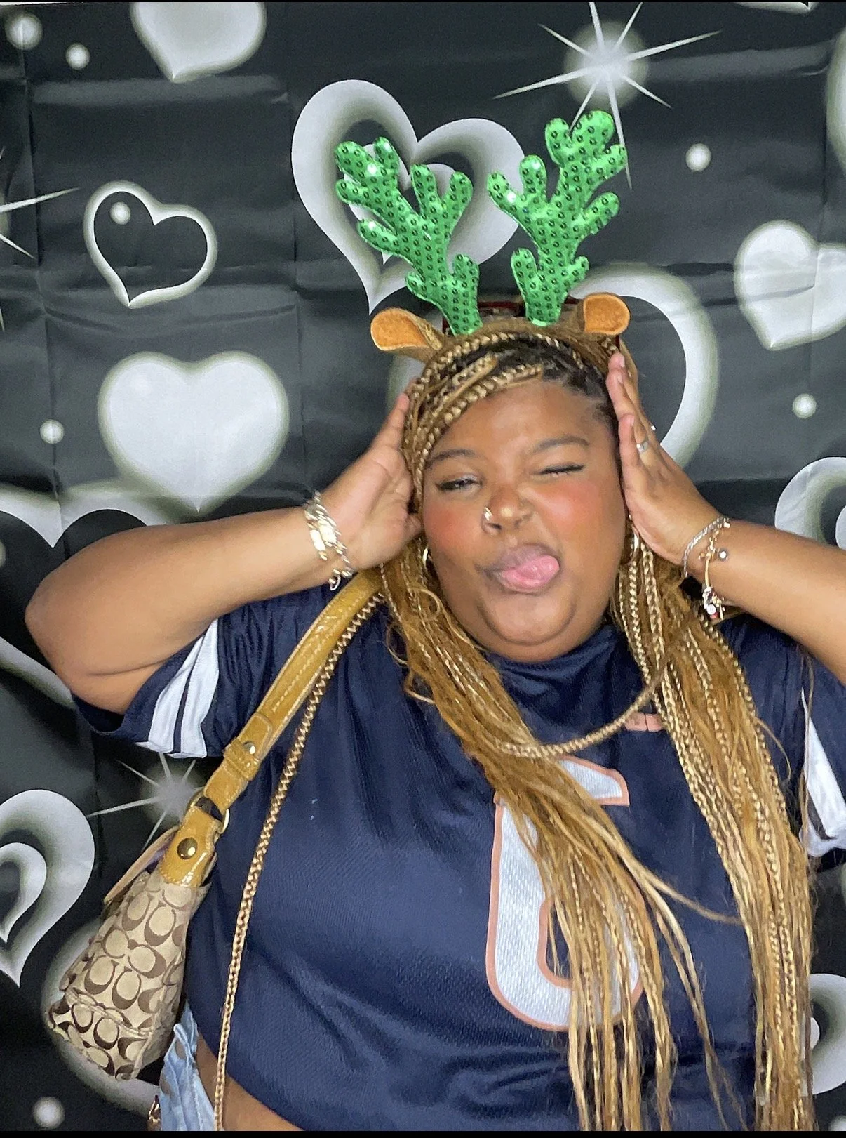 Woman with braided hair wearing reindeer antlers and green glittery antlers headband, making a fun face with her tongue out, standing in front of a black and white heart-themed backdrop.