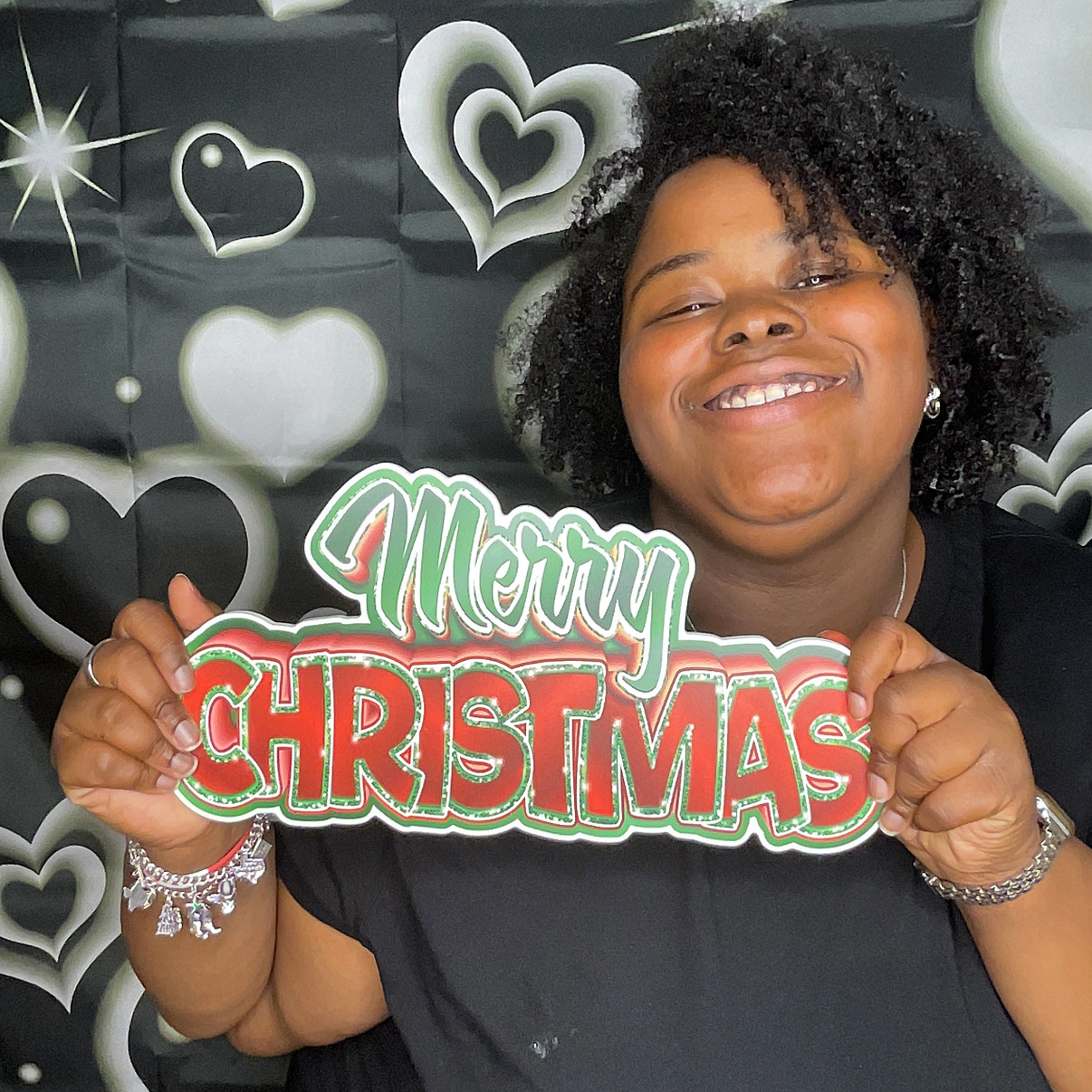 A smiling woman holding a 'Merry Christmas' sign in front of a black and white heart-patterned backdrop.