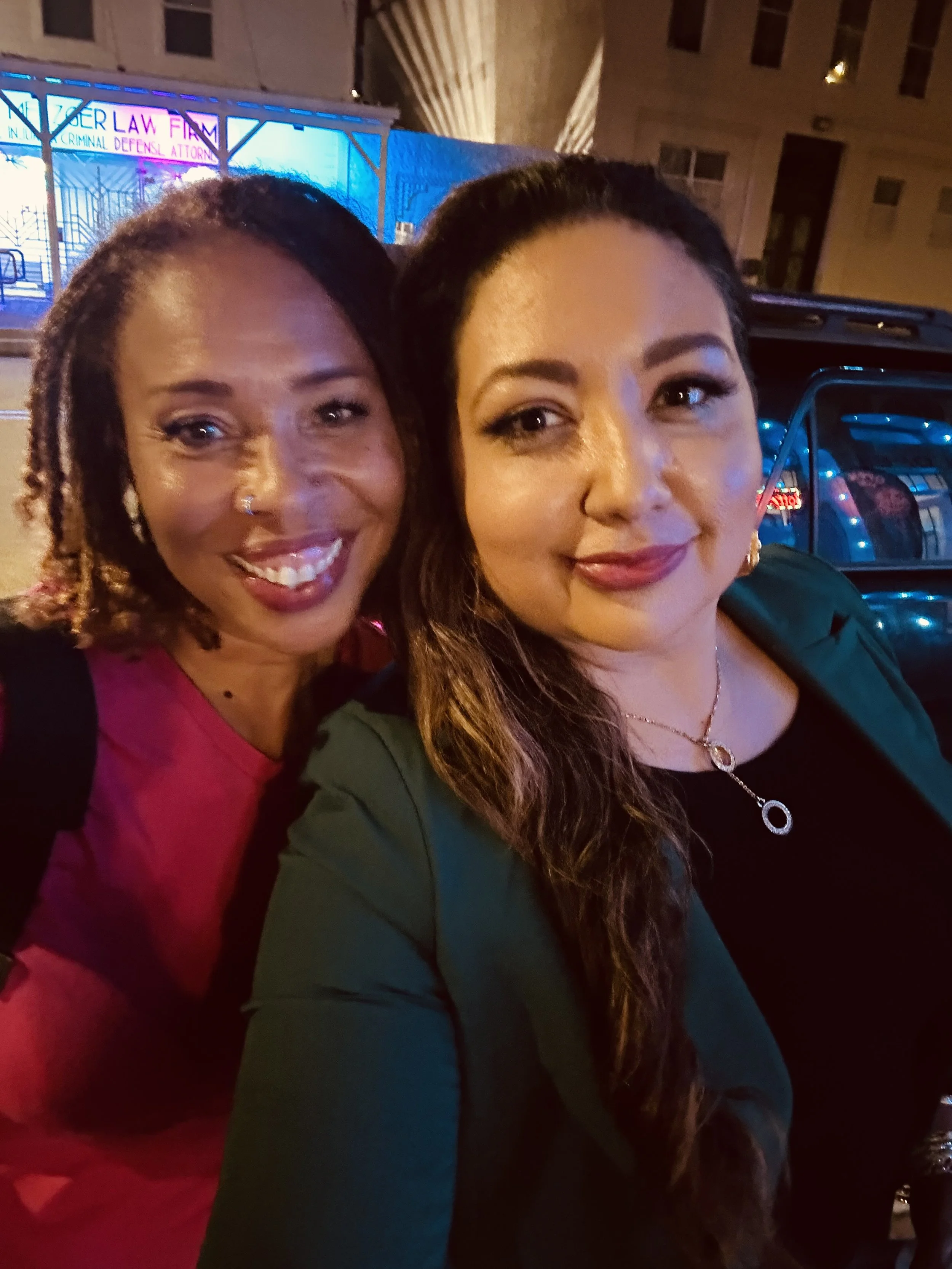 Two women taking a selfie on a city street at night, with a building and a parked car in the background.