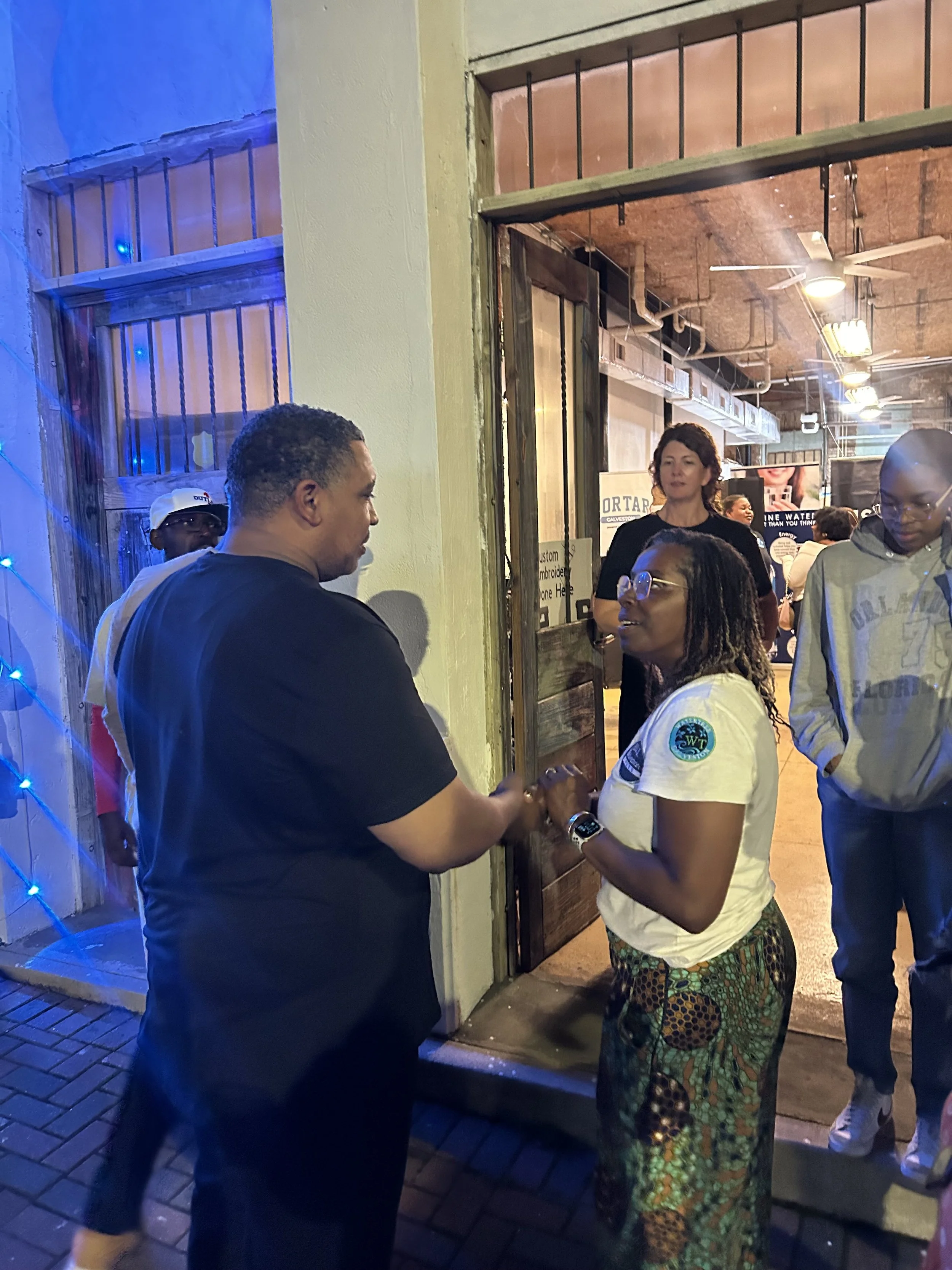 A man in a black t-shirt and a woman in a white t-shirt hold hands and look at each other outside a building, with other people in the background.