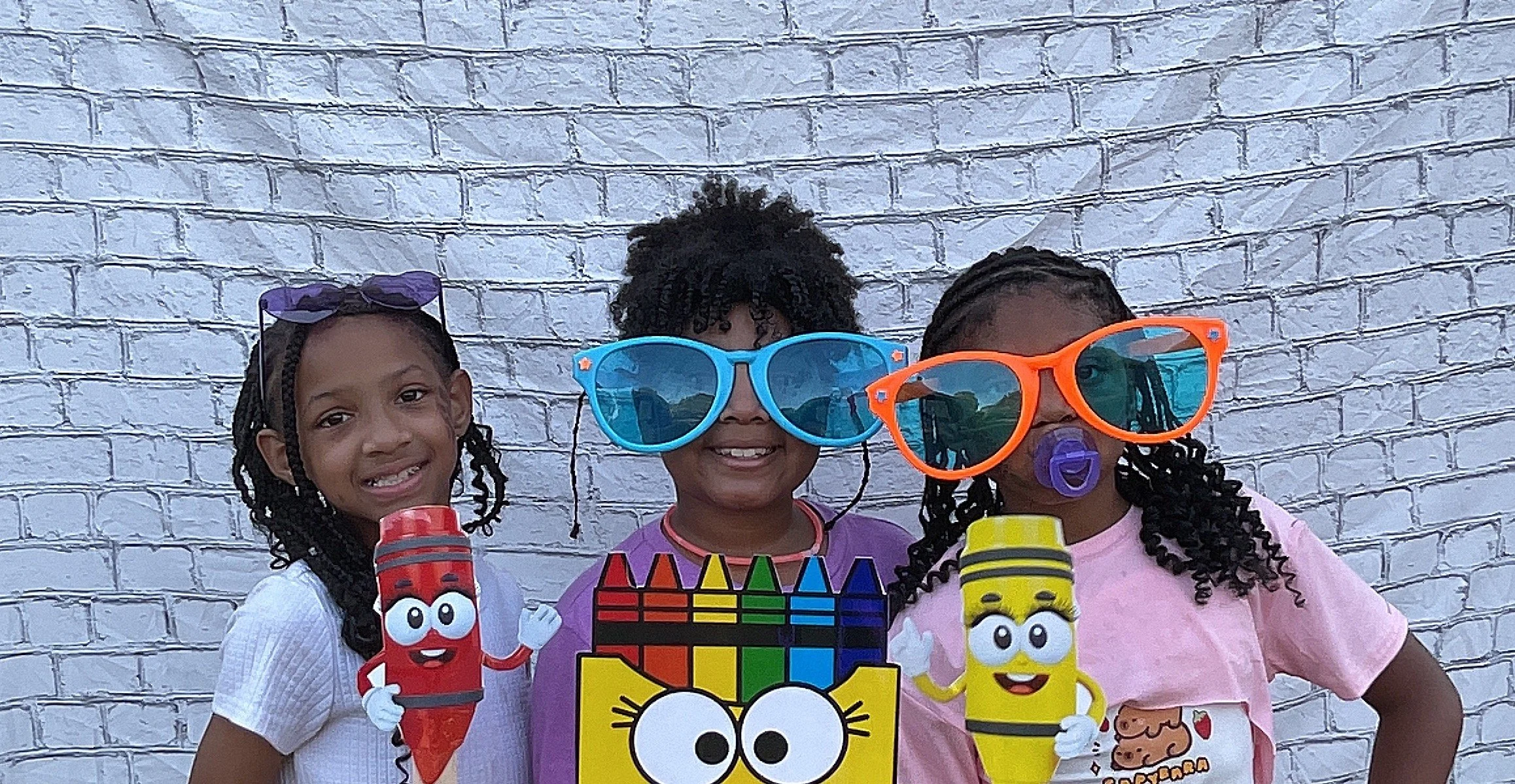 Three young girls stand against a white brick wall, wearing oversized sunglasses and holding cartoon crayon and crayon box props, smiling for the camera.