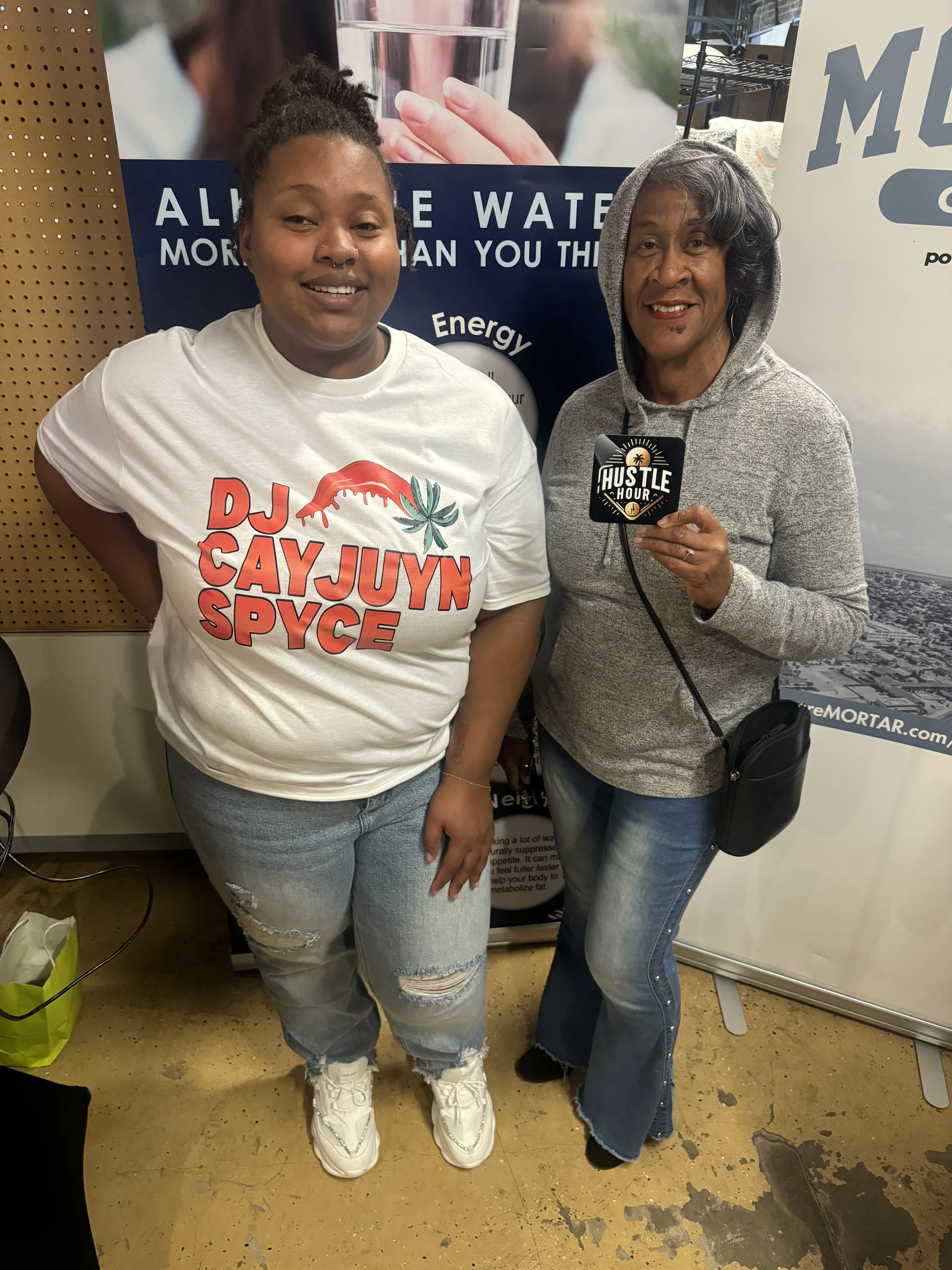 Two women standing together at a promotional event, one wearing a DJ Cay Juyn Spice T-shirt and the other holding an 'Hustle Hour' badge, with various banners and equipment in the background.