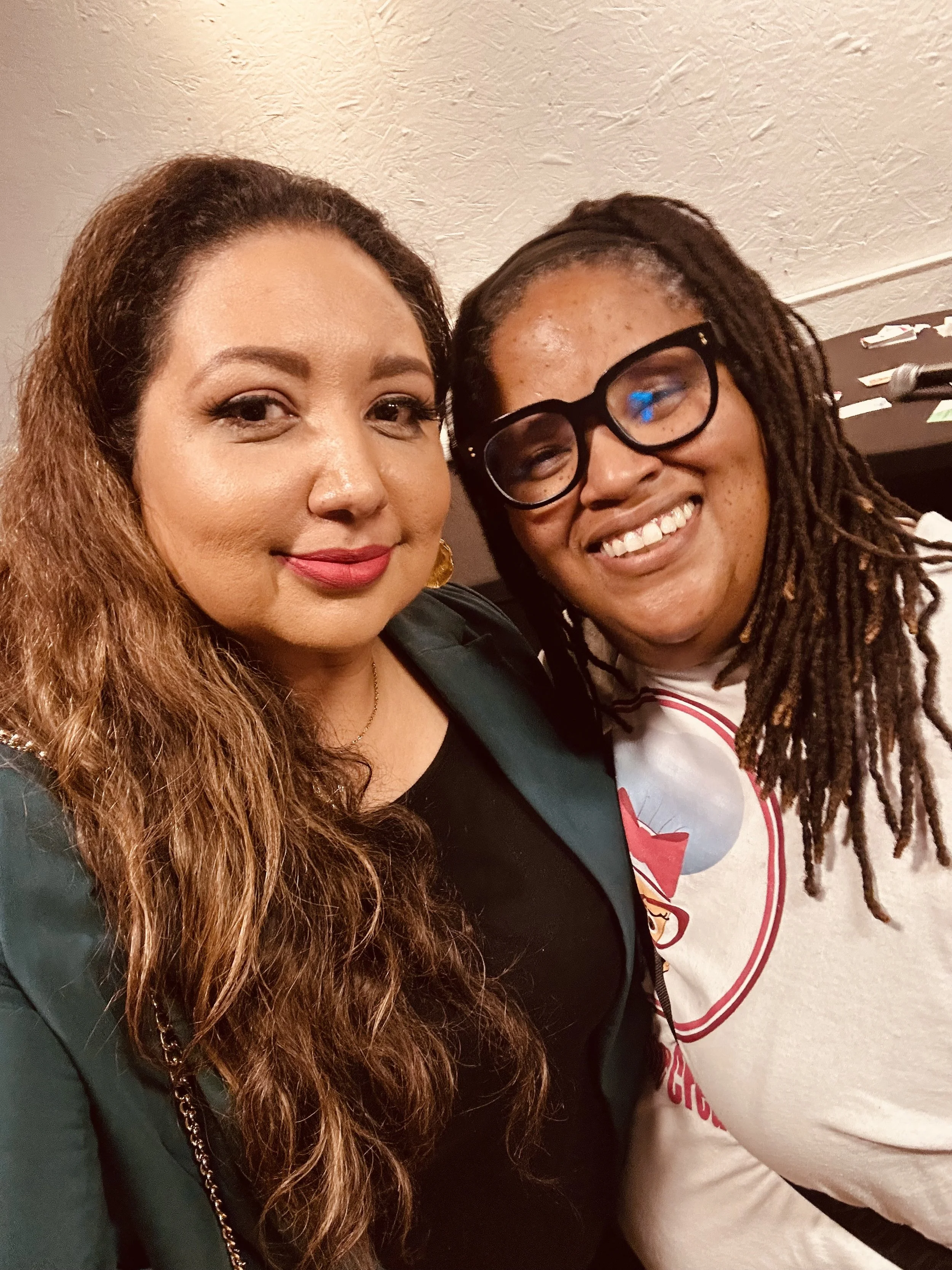 Two women smiling and taking a selfie together indoors. One has long wavy brown hair, makeup, and a dark top. The other has dreadlocks, glasses, and a white shirt with red and grey graphics.