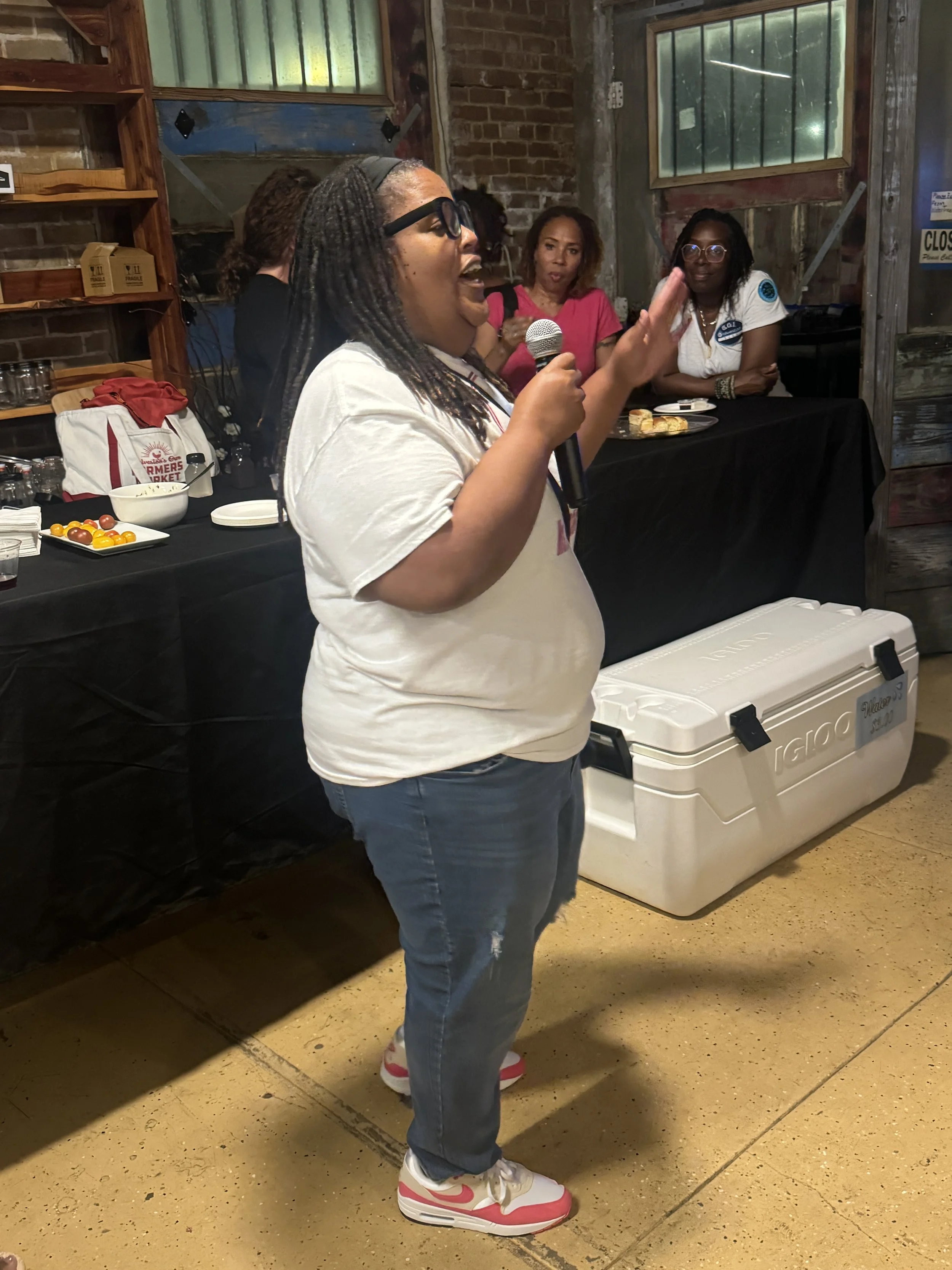 A woman speaking into a microphone at a gathering, with three women in the background standing near a table with food and drinks.