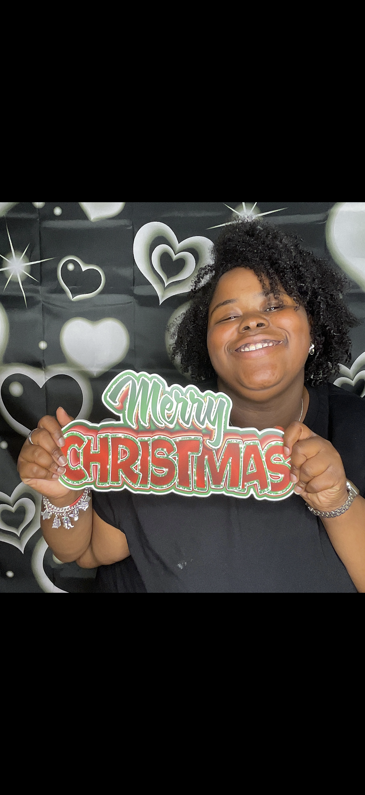A smiling woman holding a 'Merry Christmas' sign in front of a black and white heart-patterned backdrop.