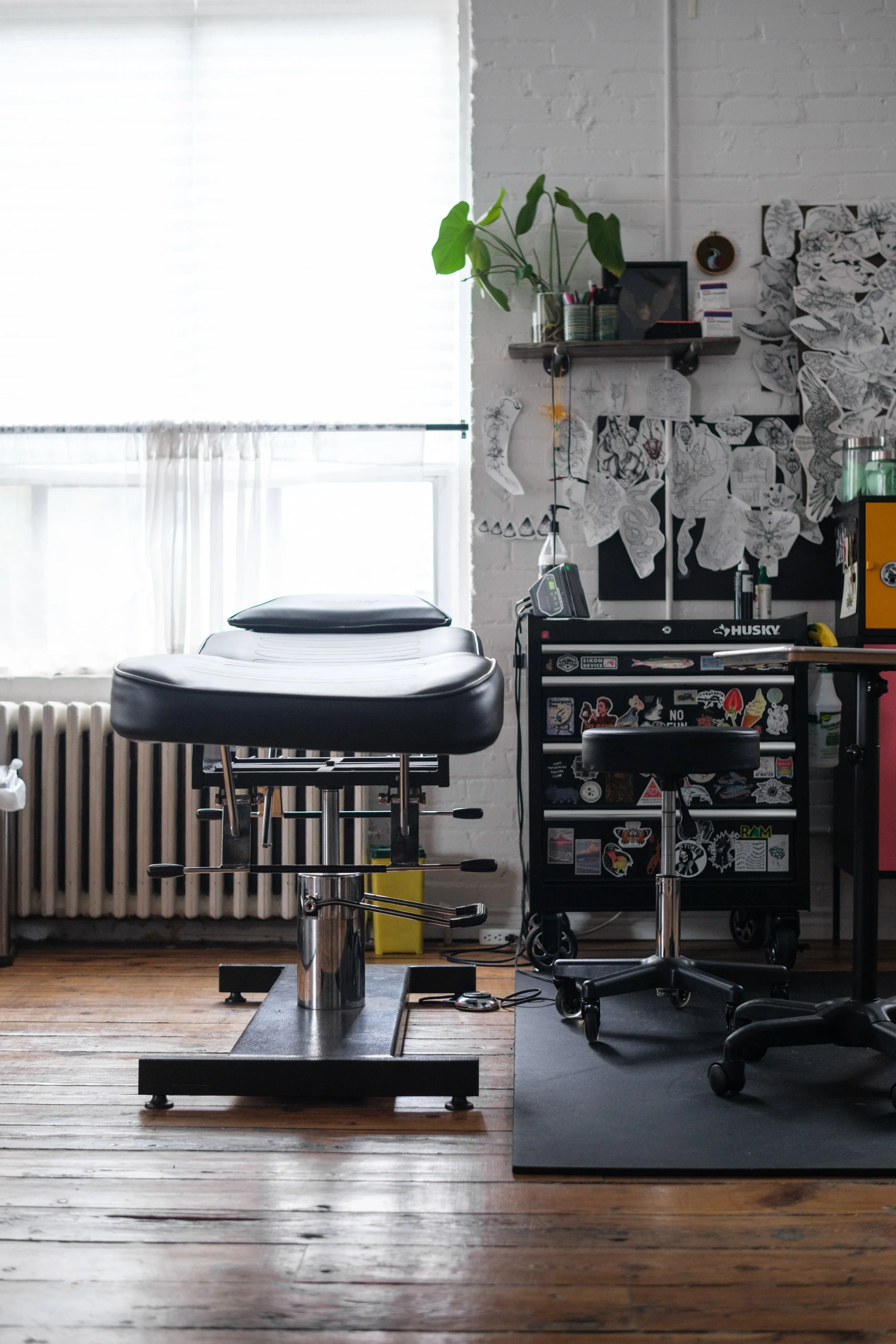 Tattoo artist's workspace with tattoo chair, rolling stool, and art supplies in an industrial-style room with wood floors and white brick walls.