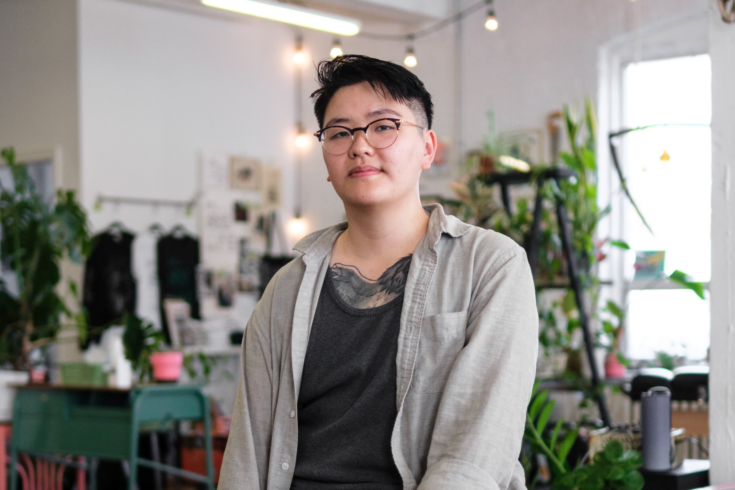 A tattoo artist with short black hair, wearing glasses, a black T-shirt, and an unbuttoned beige shirt, standing in a room filled with plants and decor, looking at the camera.