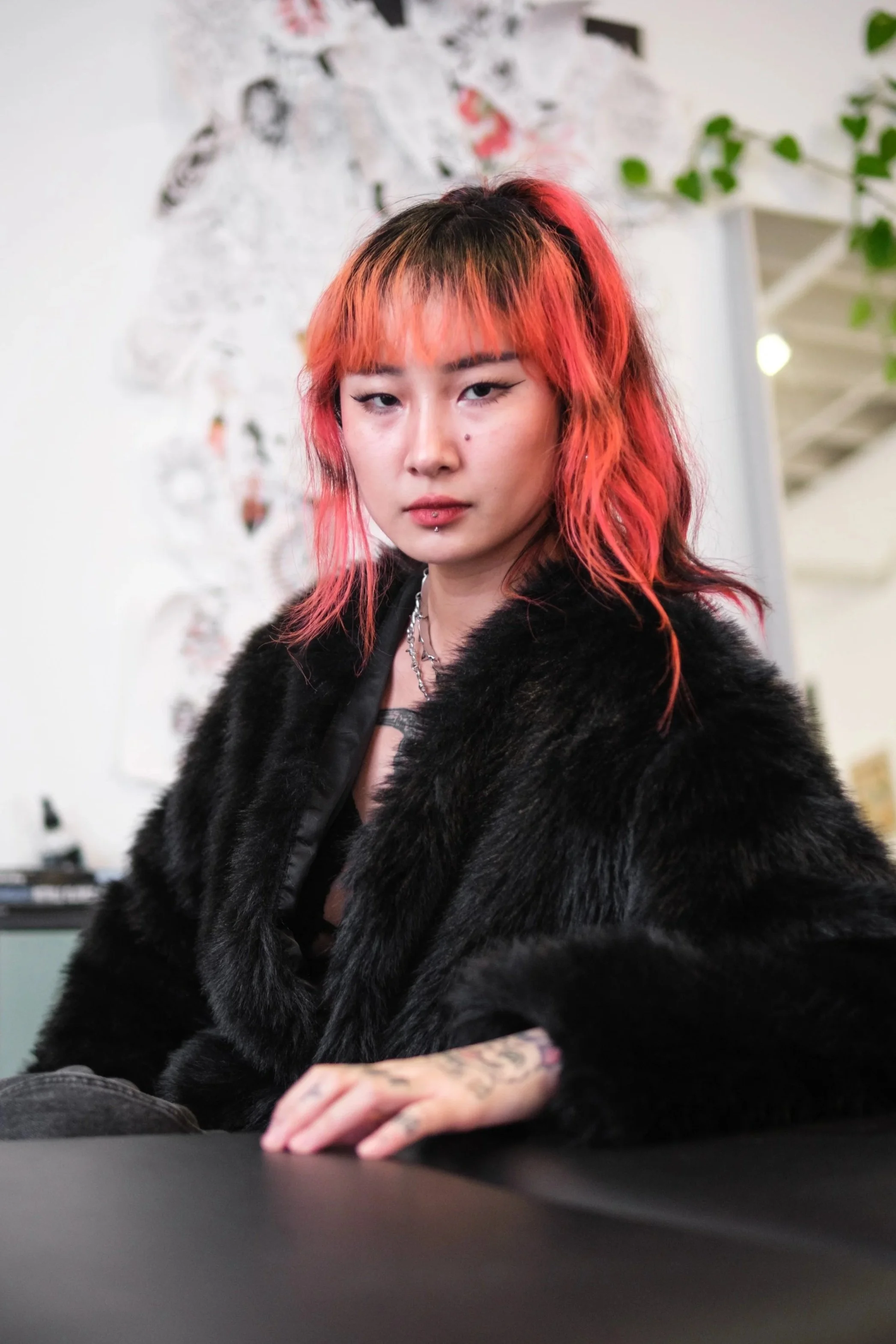 A tattoo artist with vibrant orange and black hair, wearing a black faux fur coat and silver jewelry, posing indoors with a serious expression.