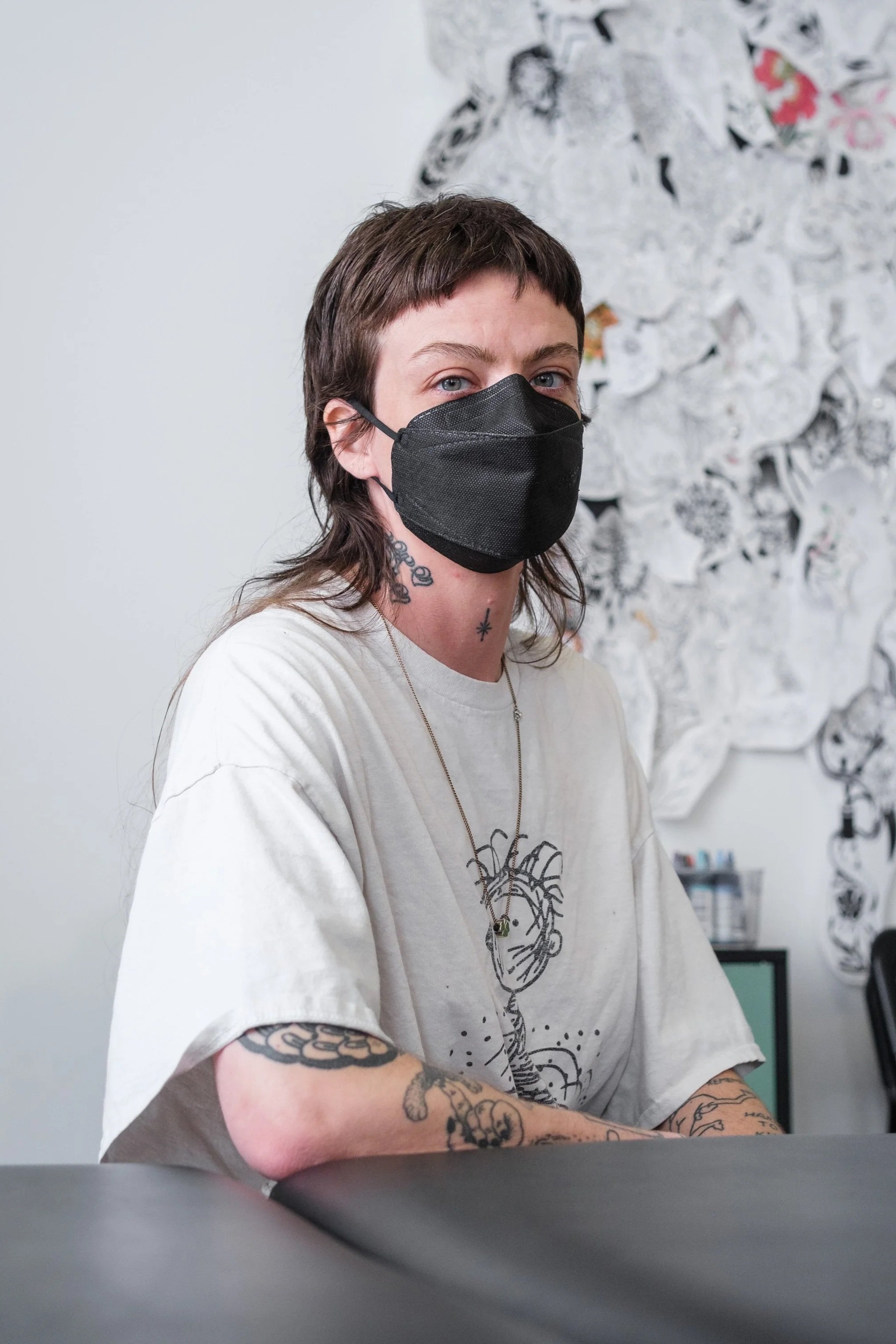 A tattoo artist with a brown mullet wearing a black face mask, tattoos on arms and neck, sitting at a table in front of abstract black and white tattoo designs.