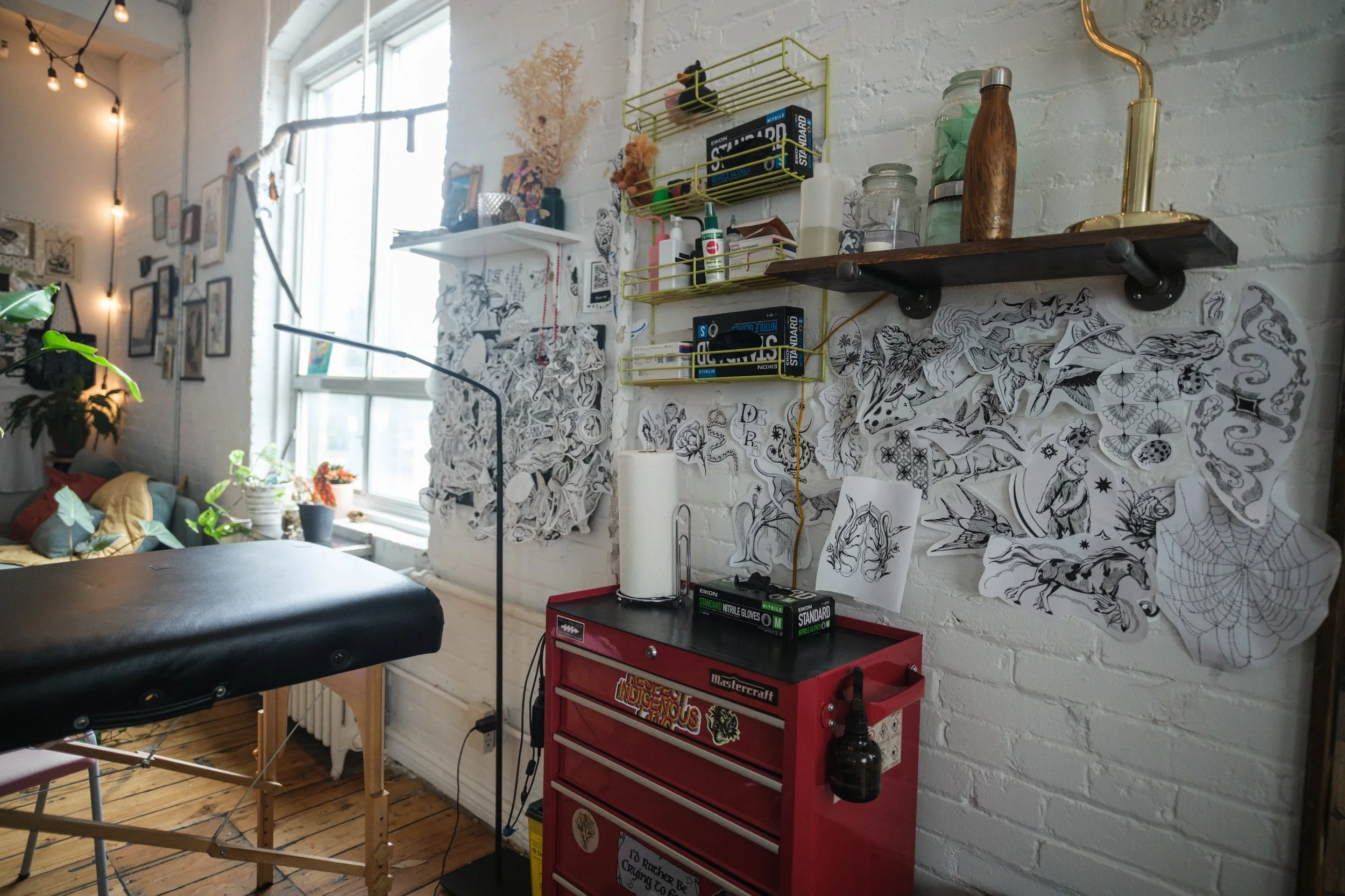 Tattoo studio with artwork sketches on white brick wall, red toolbox, massage table, and natural light from large window.