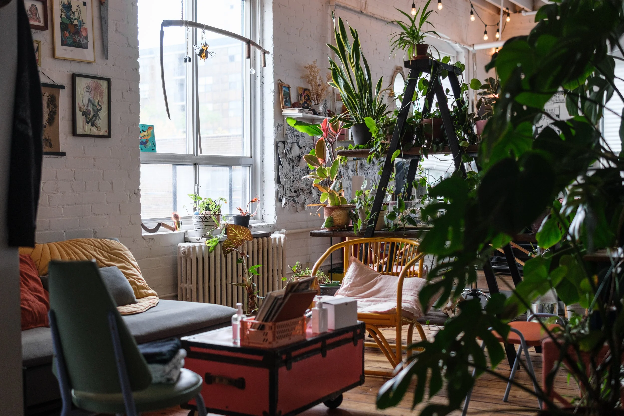 A cozy, plant-filled tattoo shop seating area with a large window, artwork on white brick walls, a gray sofa, a yellow chair, various potted plants, and a black ladder shelf with more plants. String lights are hanging from the ceiling.
