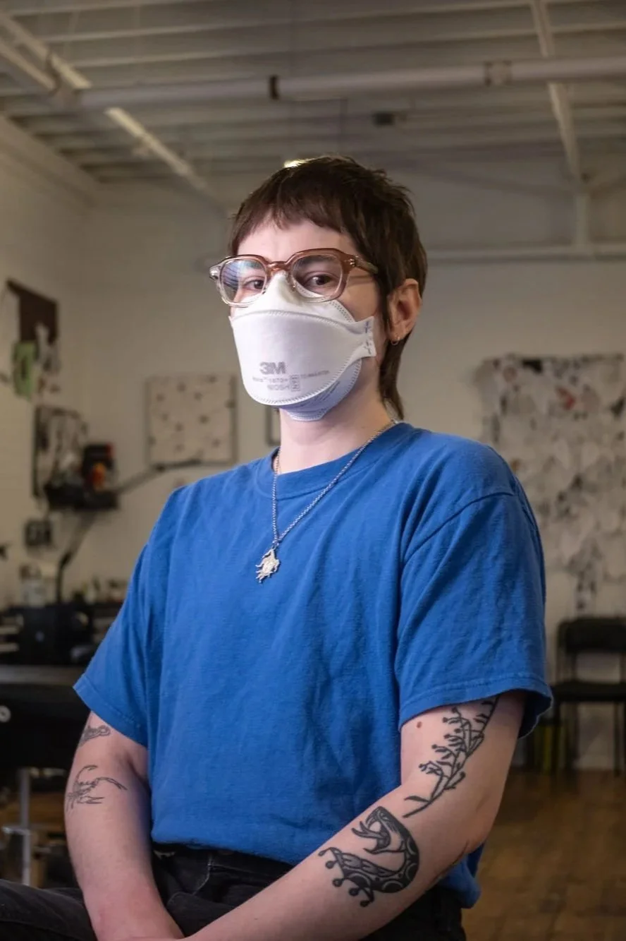 A tattoo artist wearing glasses, a face mask, a blue t-shirt, and tattoos on their arms, standing indoors.