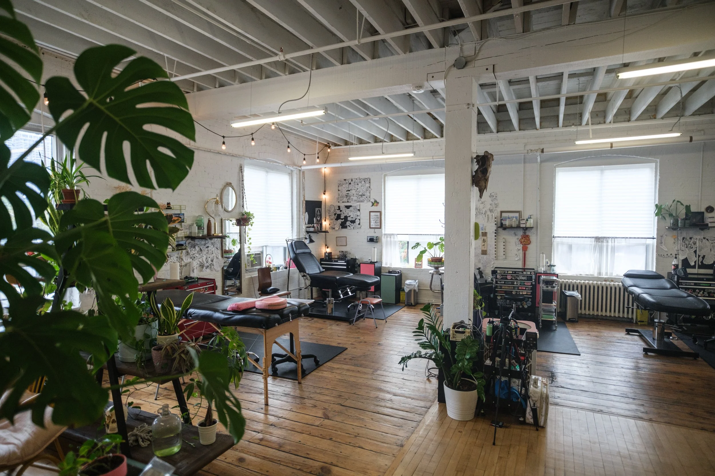 Interior view of a tattoo studio with tattoo chairs, equipment, and decor, illuminated by large windows and ceiling lights.