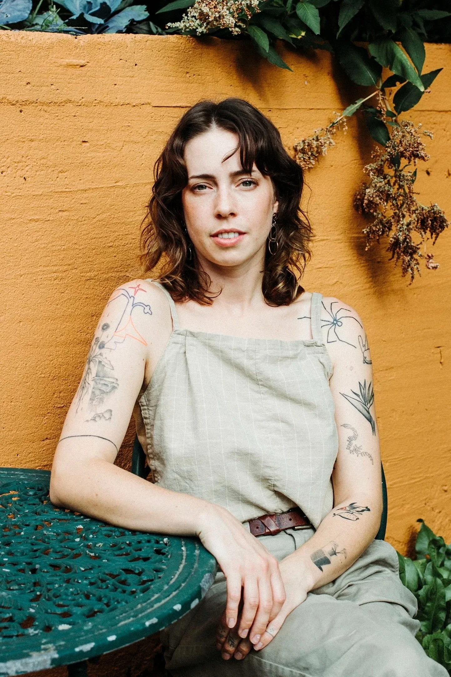 A woman with dark curly hair and tattoos on her arms, sitting outdoors against an orange wall, wearing a light-colored sleeveless top and beige pants.