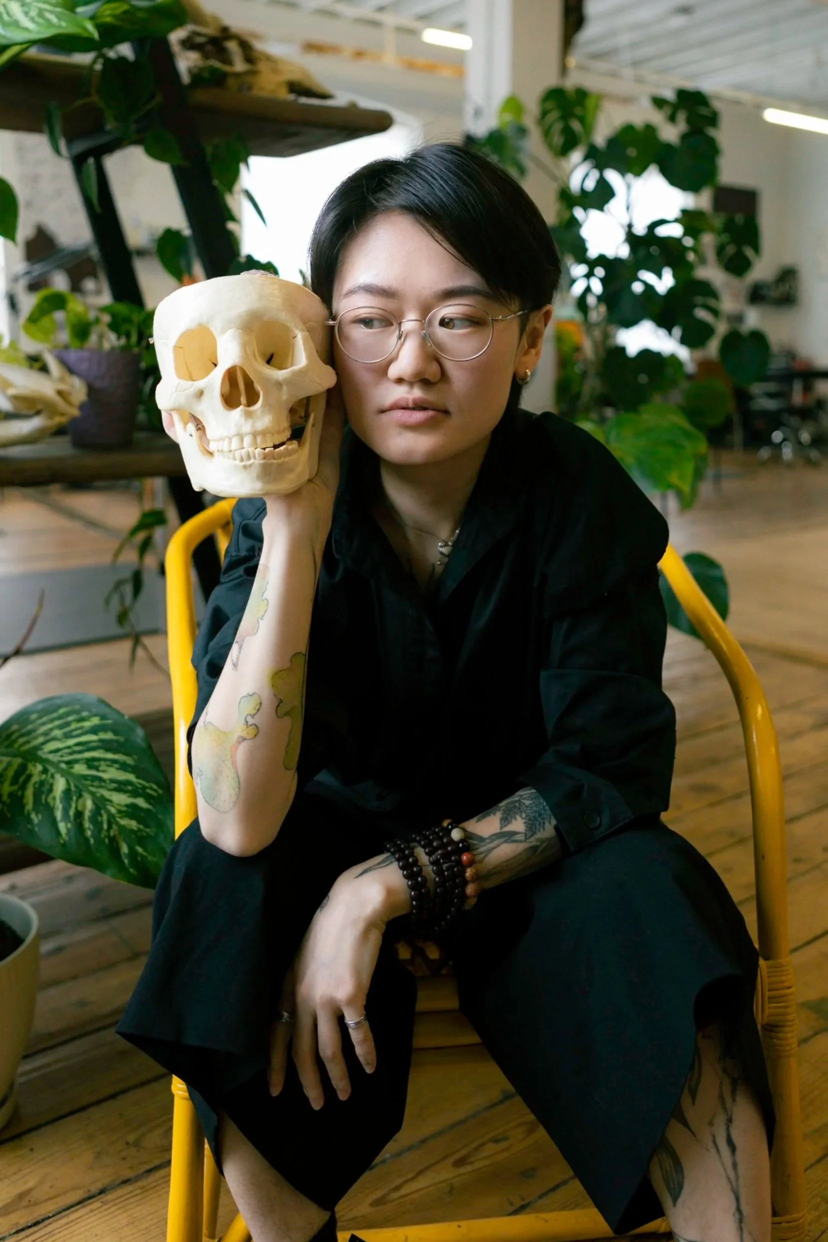 A person with short dark hair, glasses, and tattoos on their arm and leg, sitting on a yellow chair in a room with plants, holding a skull model to their face