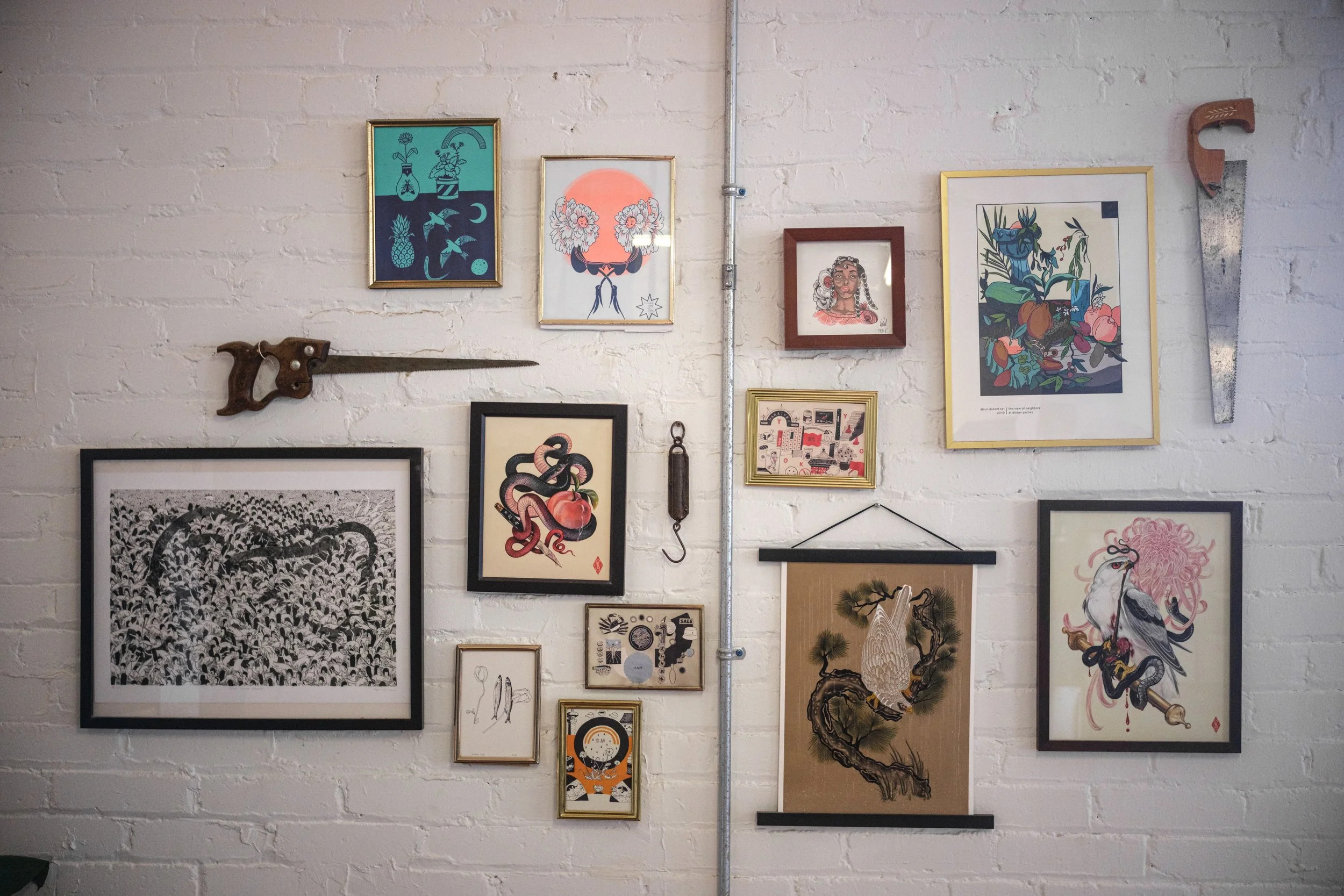 A white brick wall decorated with various framed art pieces, including illustrations, drawings, and paintings. There are vintage tools also mounted on the wall.