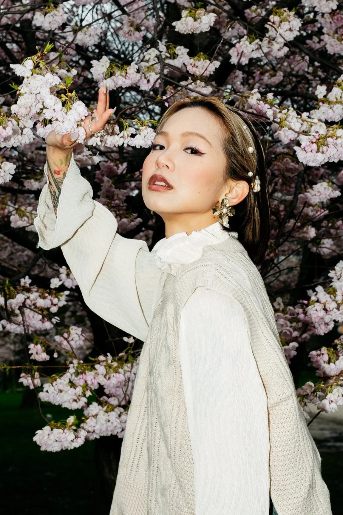 An artist with short hair and earrings standing in front of a blossoming cherry tree, touching the flowers.