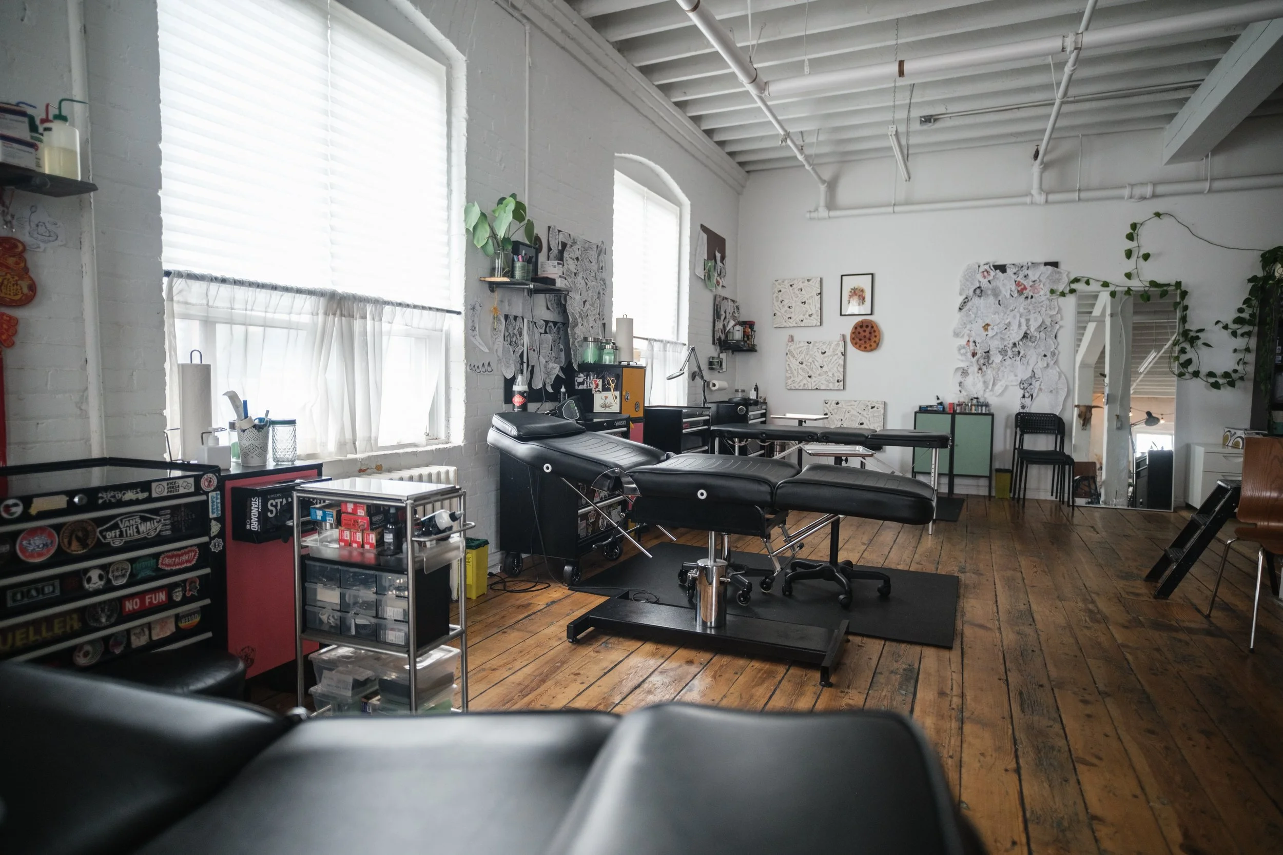 Interior view of a tattoo studio featuring a black tattoo chair, art supplies, and wall decorations, with wooden flooring and large windows.