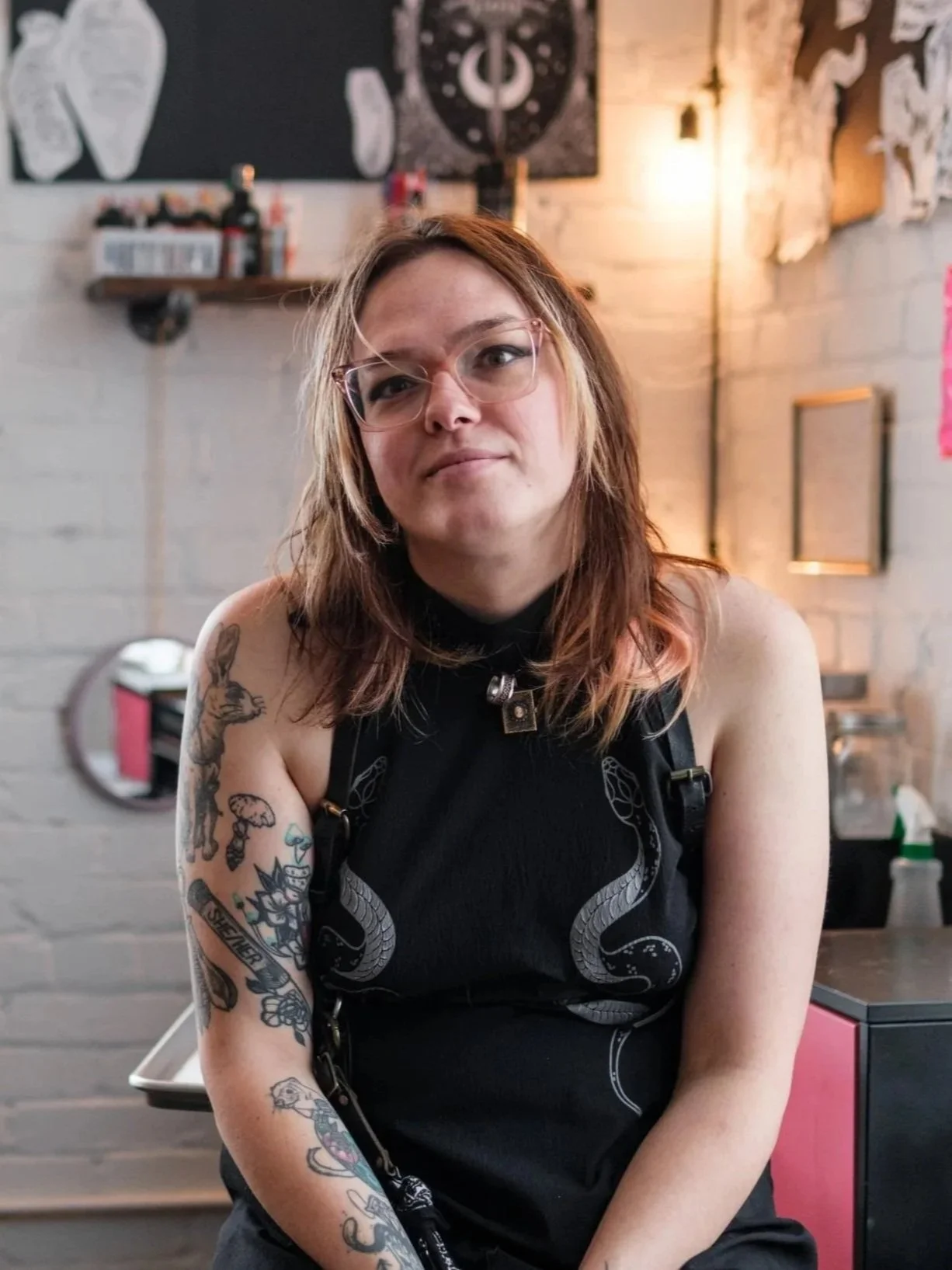 A tattoo artist with long, reddish-brown hair and glasses, wearing a black sleeveless top with snake designs, sitting inside a tattoo shop with tattoo artwork on the wall behind her.