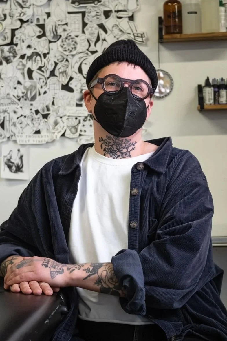 A tattoo artis sitting at a table with arms crossed. They are wearing a dark jacket over a white shirt, and have tattoos on their neck and hands. The background includes black and white artwork on the wall and shelves with bottles and containers.