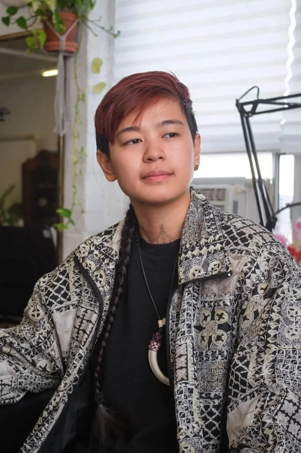 A tattoo artist with short hair and a braid, wearing a patterned jacket and a necklace with a pendant, sitting indoors near a window with white blinds and greenery in the background.