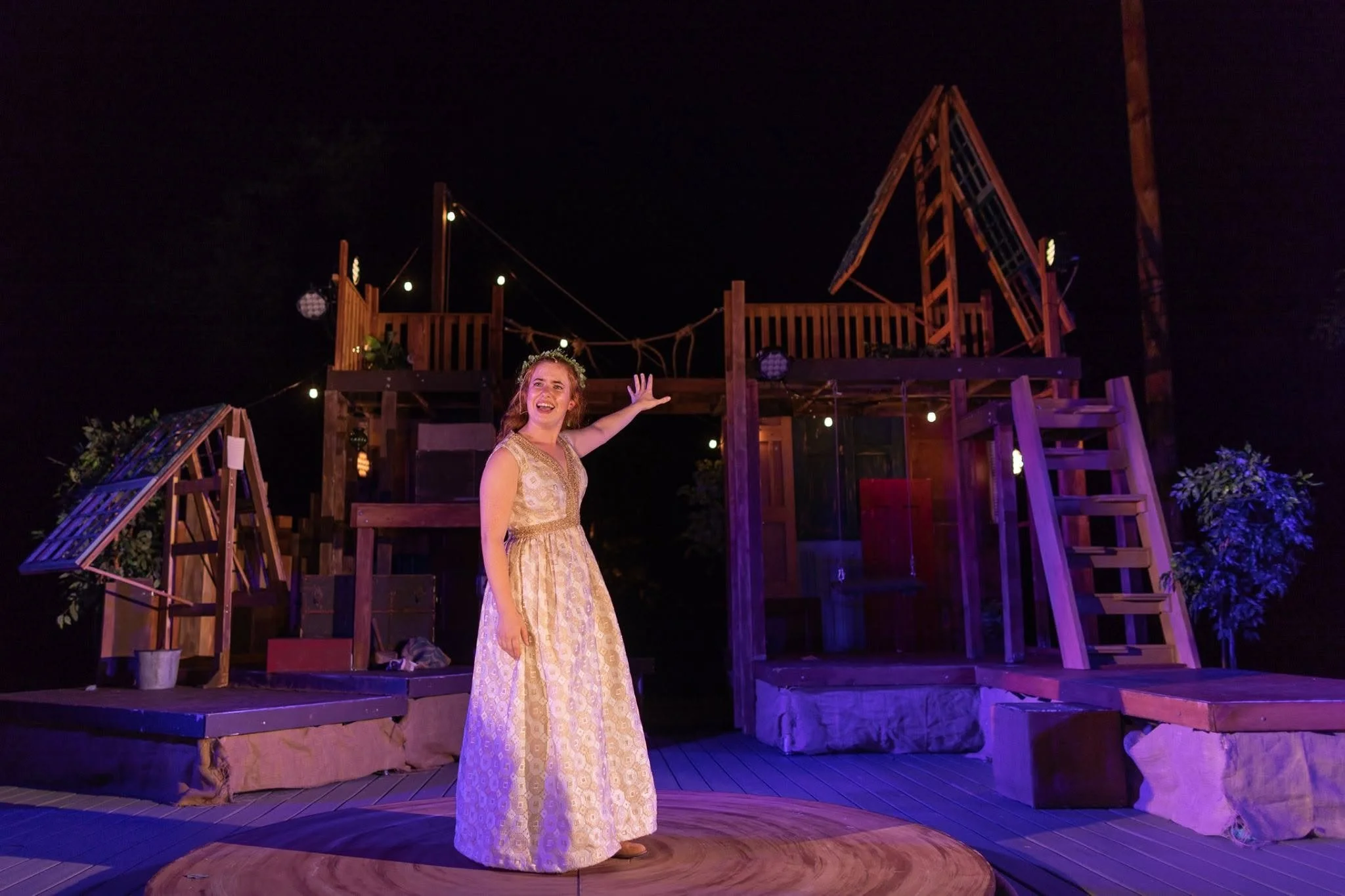 A woman in a cream-colored dress with gold accents standing on a wooden stage at night, smiling and gesturing with her right hand. The stage features wooden structures, plants, and hanging lights, with a dark sky in the background.