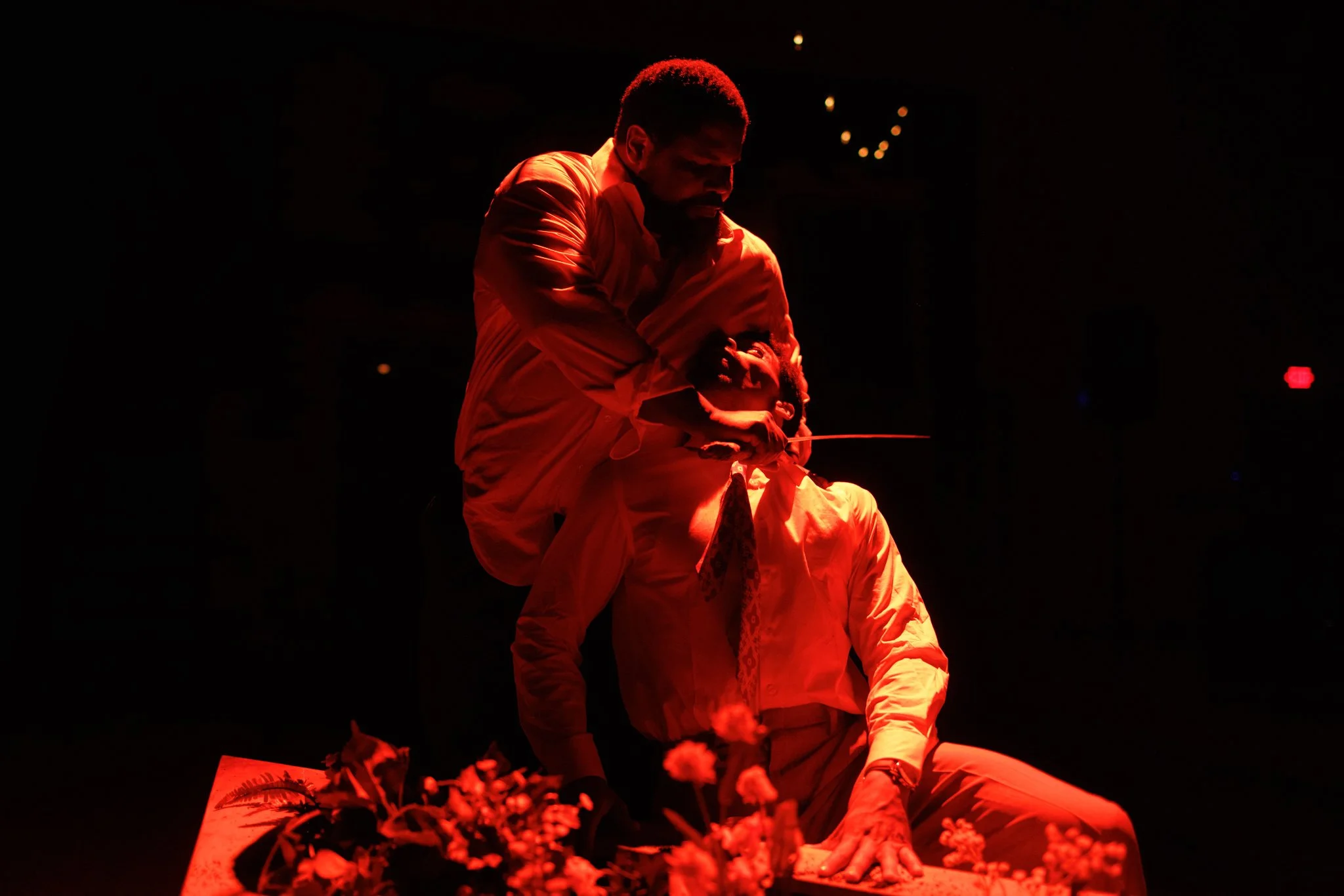A man with a theatrical knife on another man's neck in a dimly lit setting with red lighting.