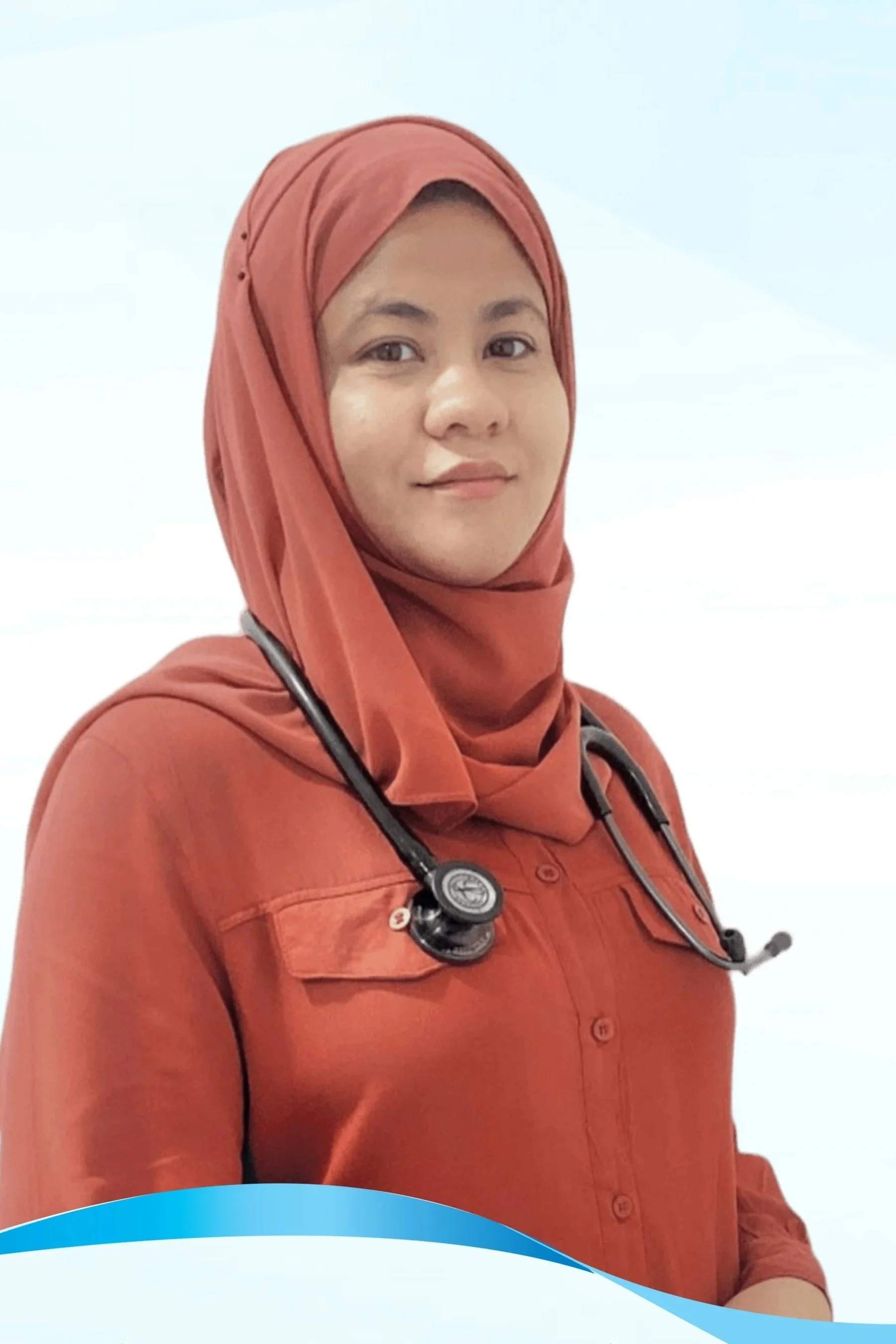 A young woman wearing a rust-colored hijab and shirt, with a stethoscope around her neck, standing indoors.