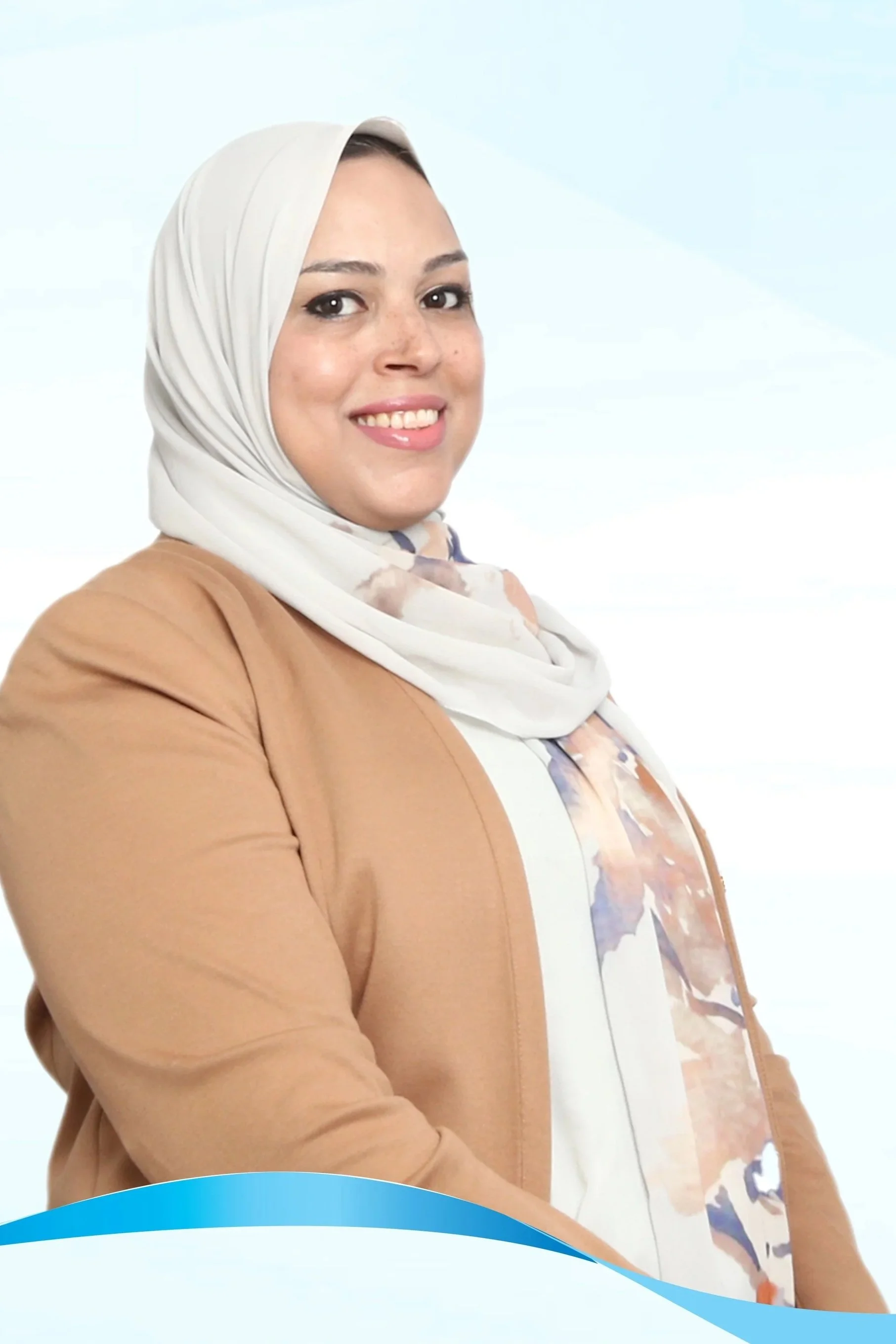A woman wearing a white and pink patterned hijab and a beige blazer, smiling.