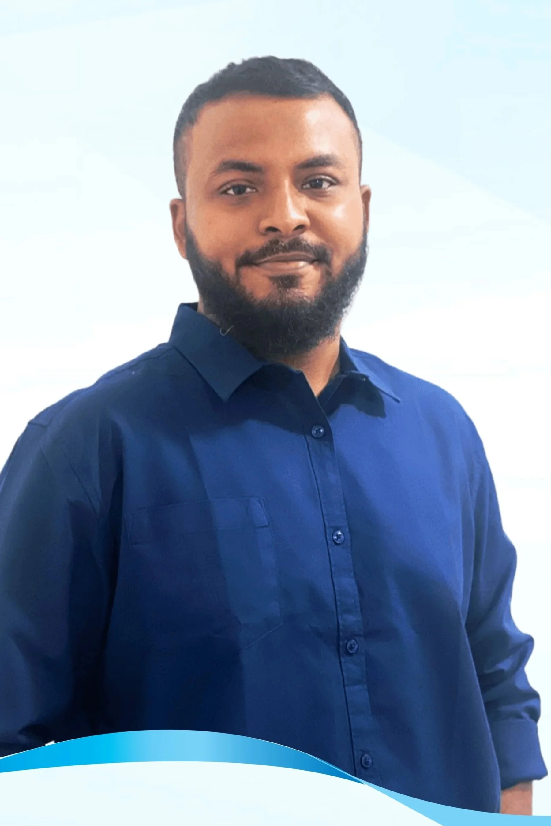 A man with a trimmed beard and short hair wearing a navy blue dress shirt, standing indoors with a blurred background.