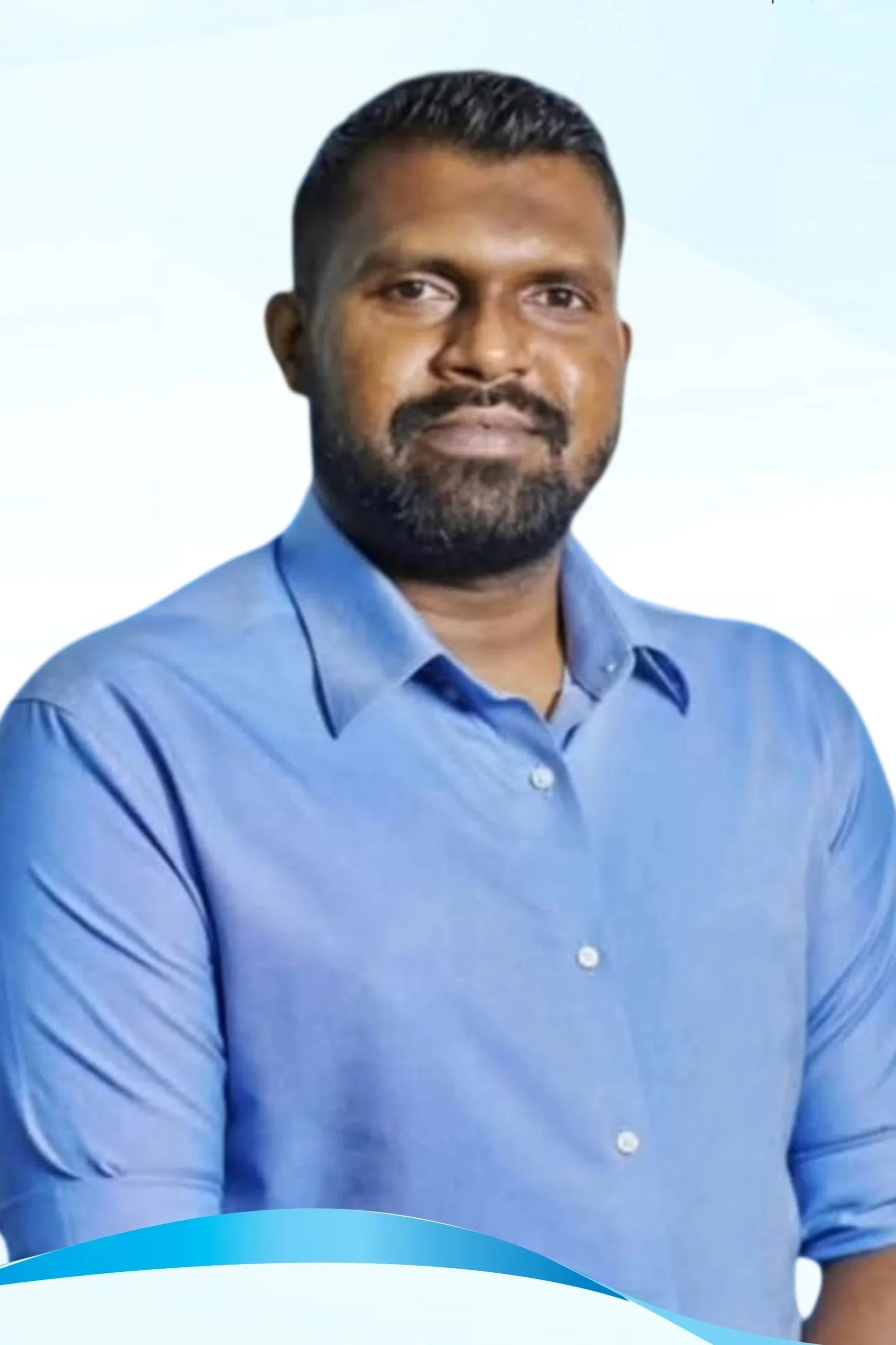 Portrait of a man with dark hair and a beard, wearing a blue collared shirt, standing in front of a blurred background.