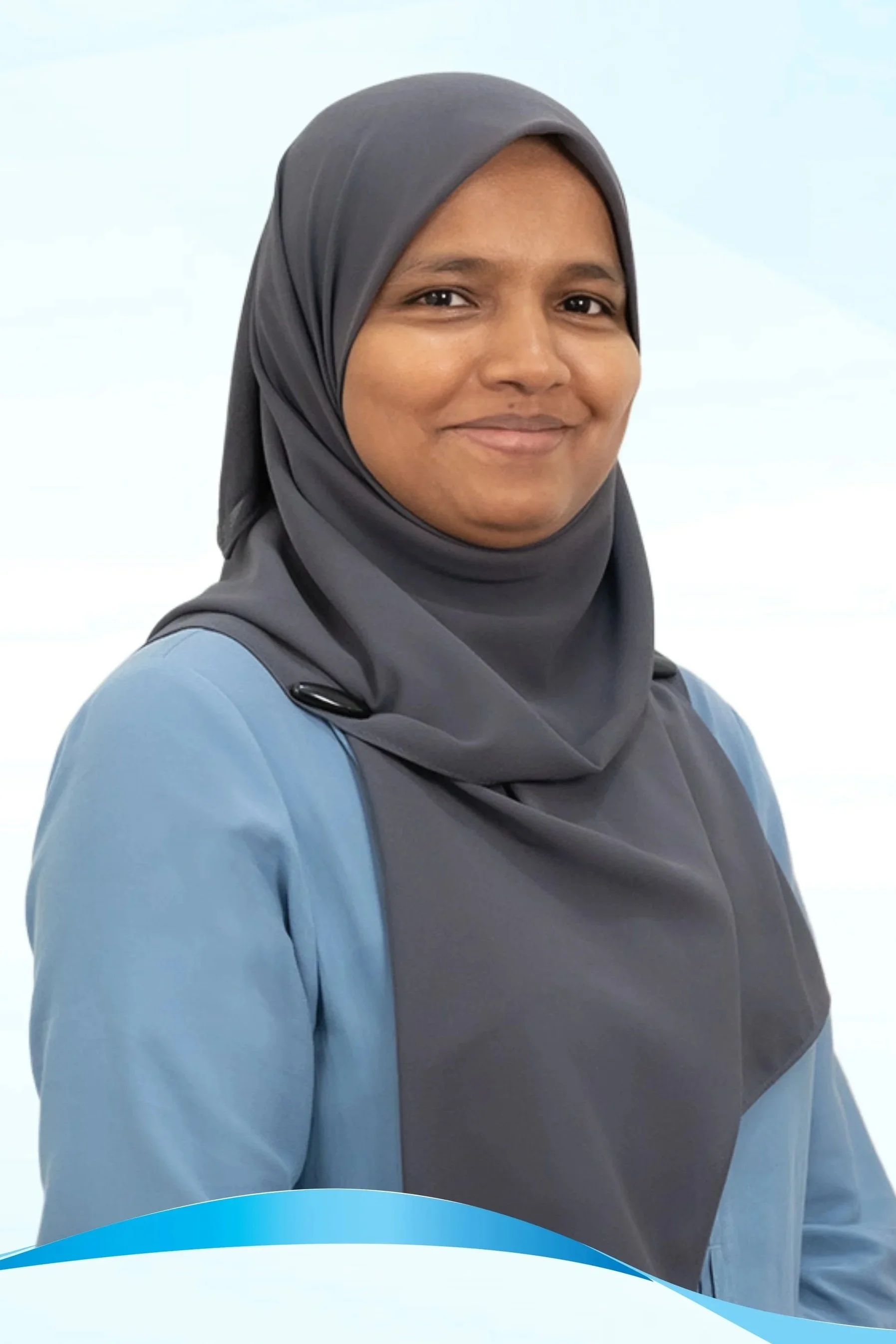A woman in a gray hijab and light blue medical uniform smiling in a healthcare setting.
