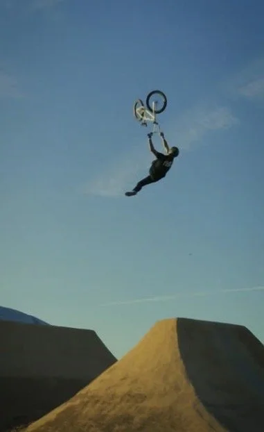 Bmx bicycle rider doing a trick over a big dirt jump for the crowd on a sunny day in front of a football stadium in the parking lot.