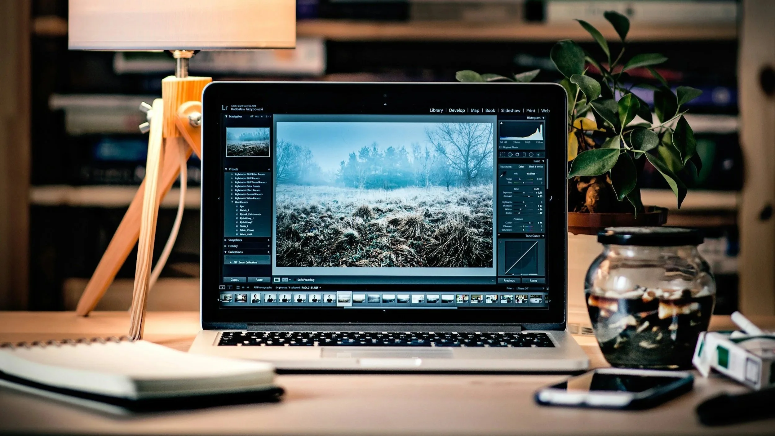 Un écran d'ordinateur portable affichant une photo de paysage enneigé, posé sur un bureau avec une pile de livres, une plante en pot, un smartphone, un téléphone portable et une lampe de bureau en bois.